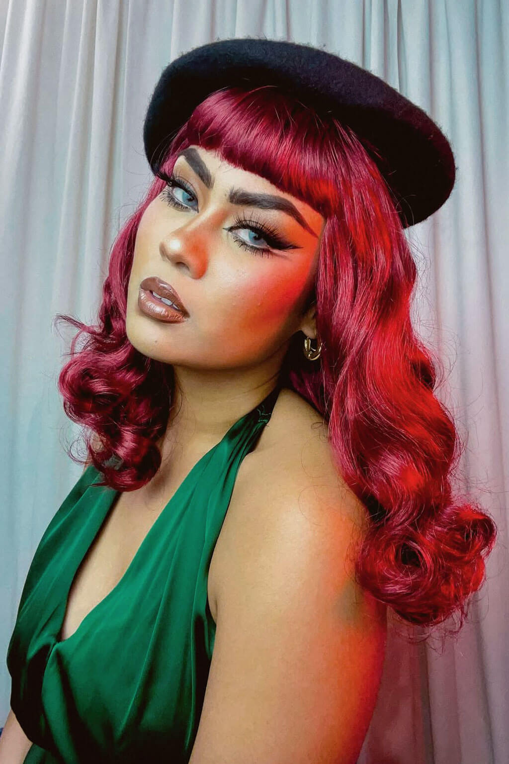 Woman with vibrant red hair and a black hat against a light curtain background. Dark cherry red 1950s pinup style wig: Sandra