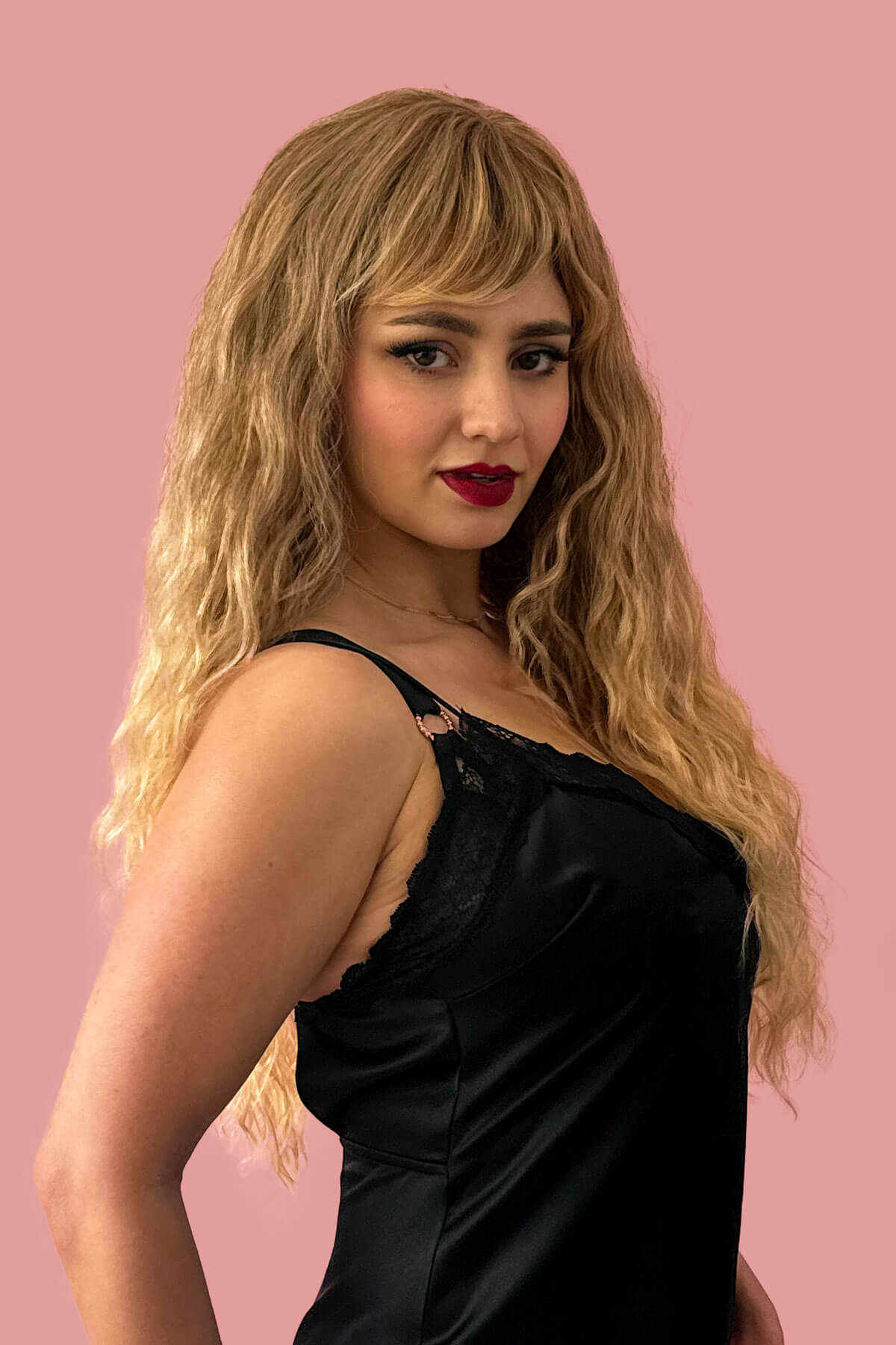 Woman with blonde hair and red lipstick wearing a black dress against a pink background. Long brown crimped wig, brown and blonde dip dye (ombre): Tiggy