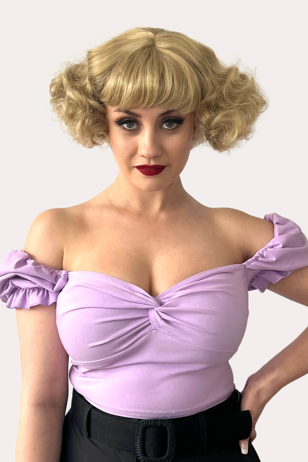 Woman wearing a purple off-shoulder top with a plain background. A short blonde vintage style wig with waves: Joy