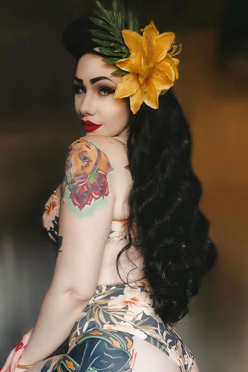 Woman with tattoos and a yellow flower in her hair against a dark background. Long black pinup style wig with vintage hollywood waves: Veronica