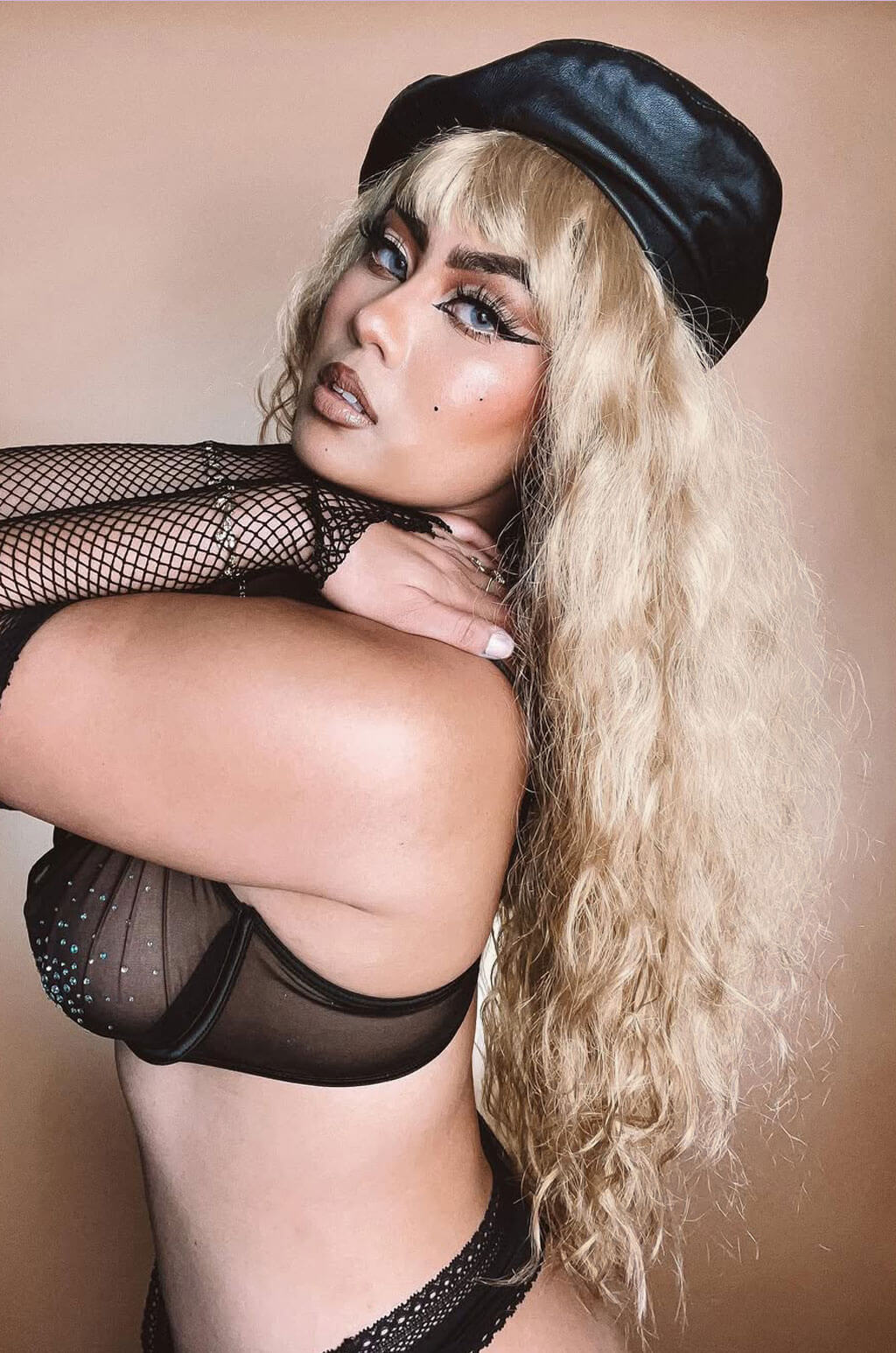 Woman wearing a black cap and fishnet top against a beige background. Long wig with crimped mermaid waves: Astrid
