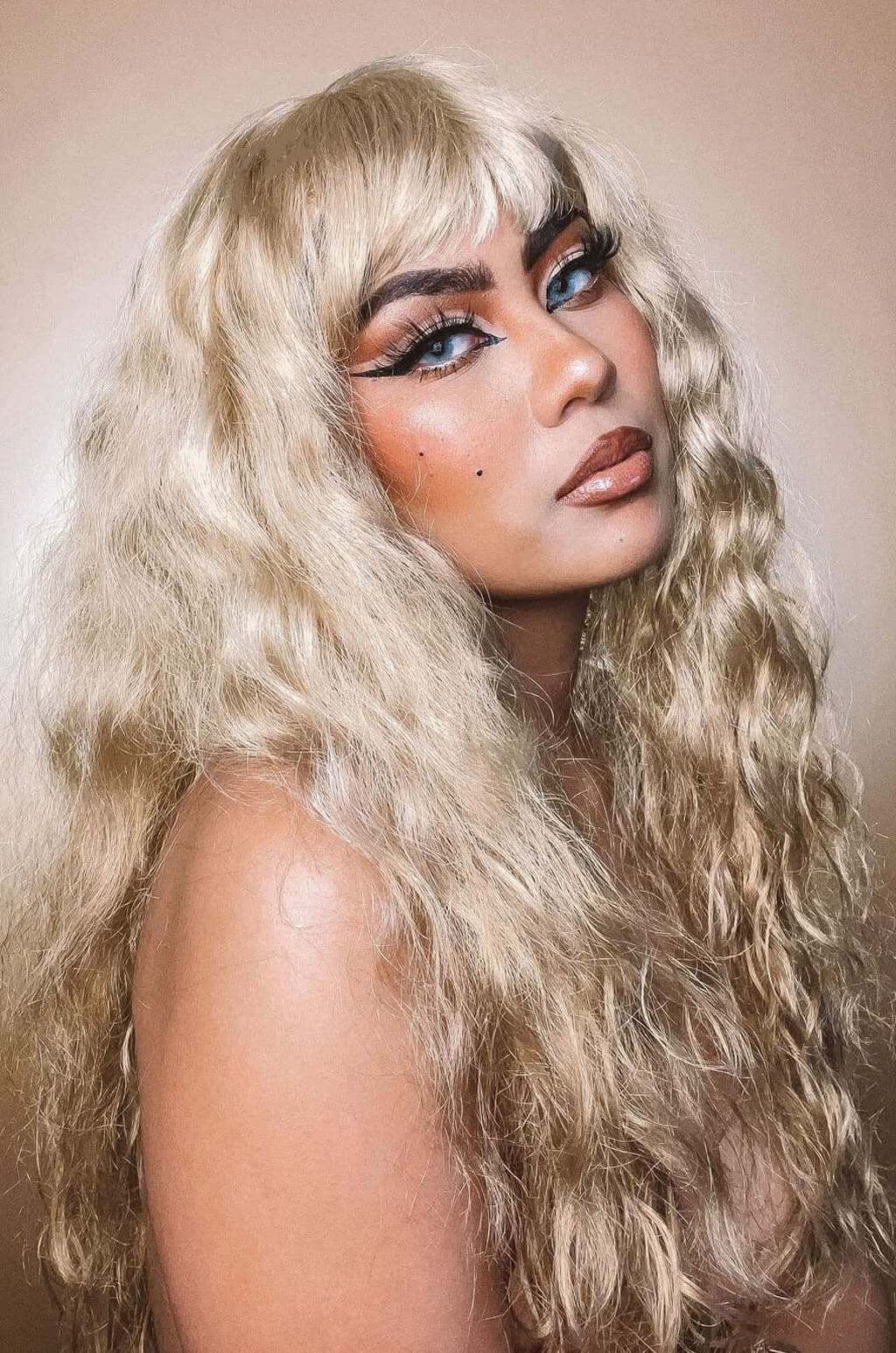 Person with long blonde hair and bold makeup against a neutral background. Long wig with crimped mermaid waves: Astrid