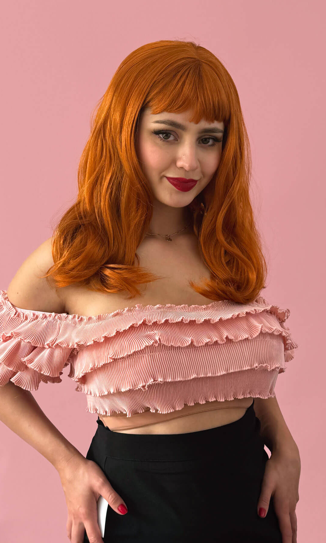 Person with ginger hair wearing a pink ruffled top against a pink background. Ginger long wavy bob (LOB) wig with straight retro fringe: Cerissa