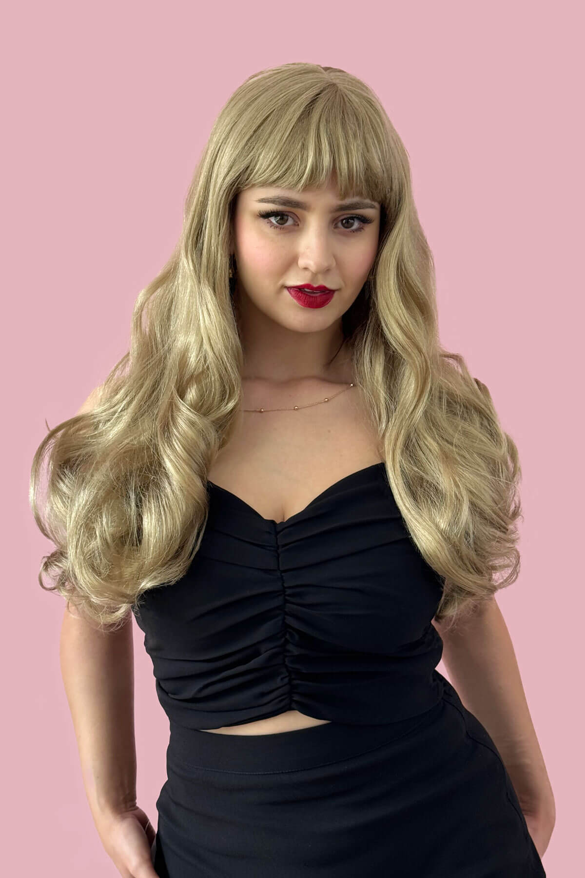 Woman with long blonde wig and black dress against a pink background. Long blonde wavy wig with pinup-style fringe: Elena