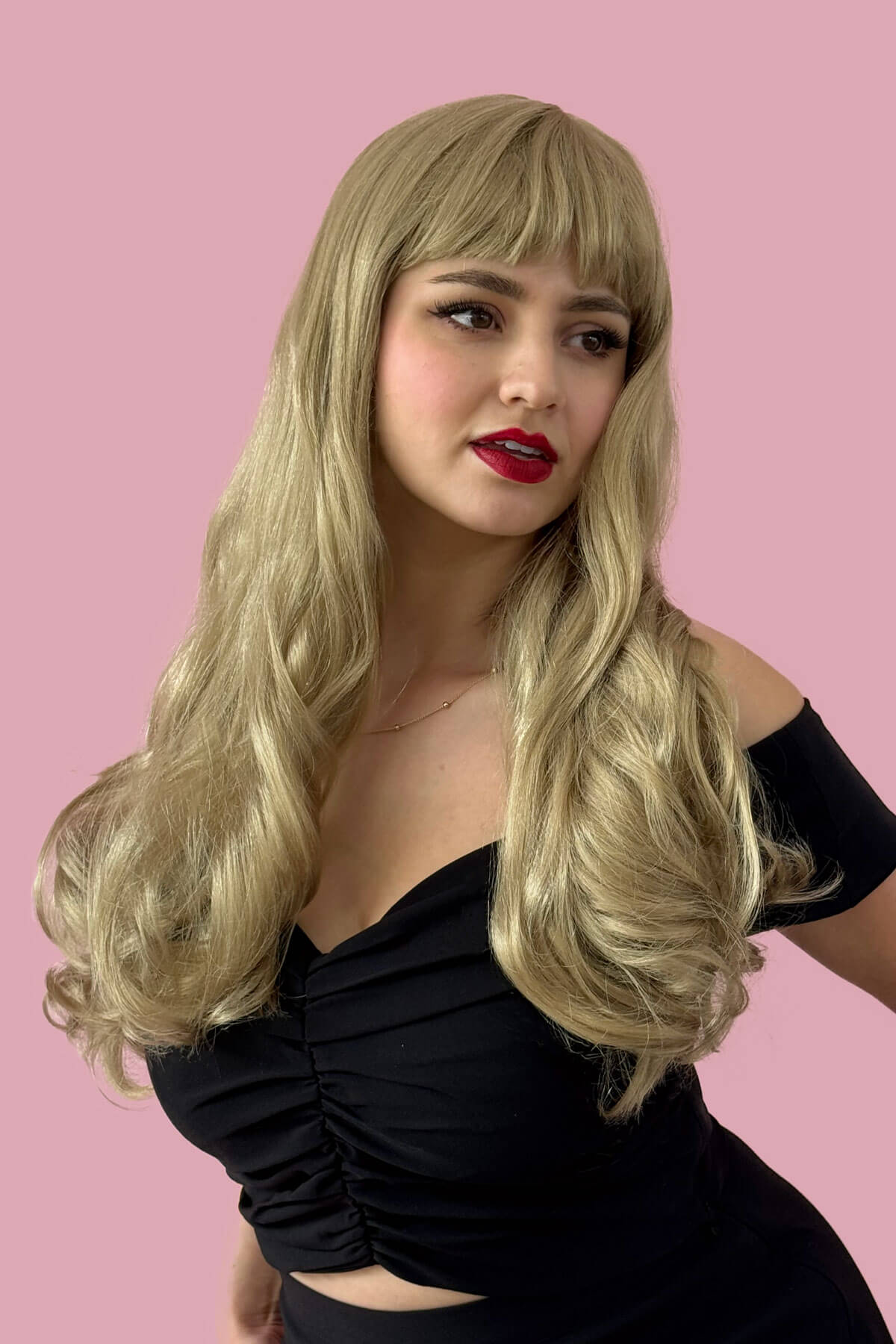 Woman with long blonde wig wearing a black top against a pink background. Long blonde wavy wig with pinup-style fringe: Elena