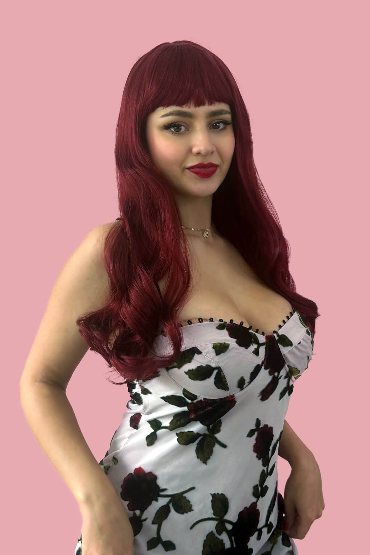 Woman with red hair wearing a floral dress against a pink background. Long red wig with straight retro-style fringe: Esther
