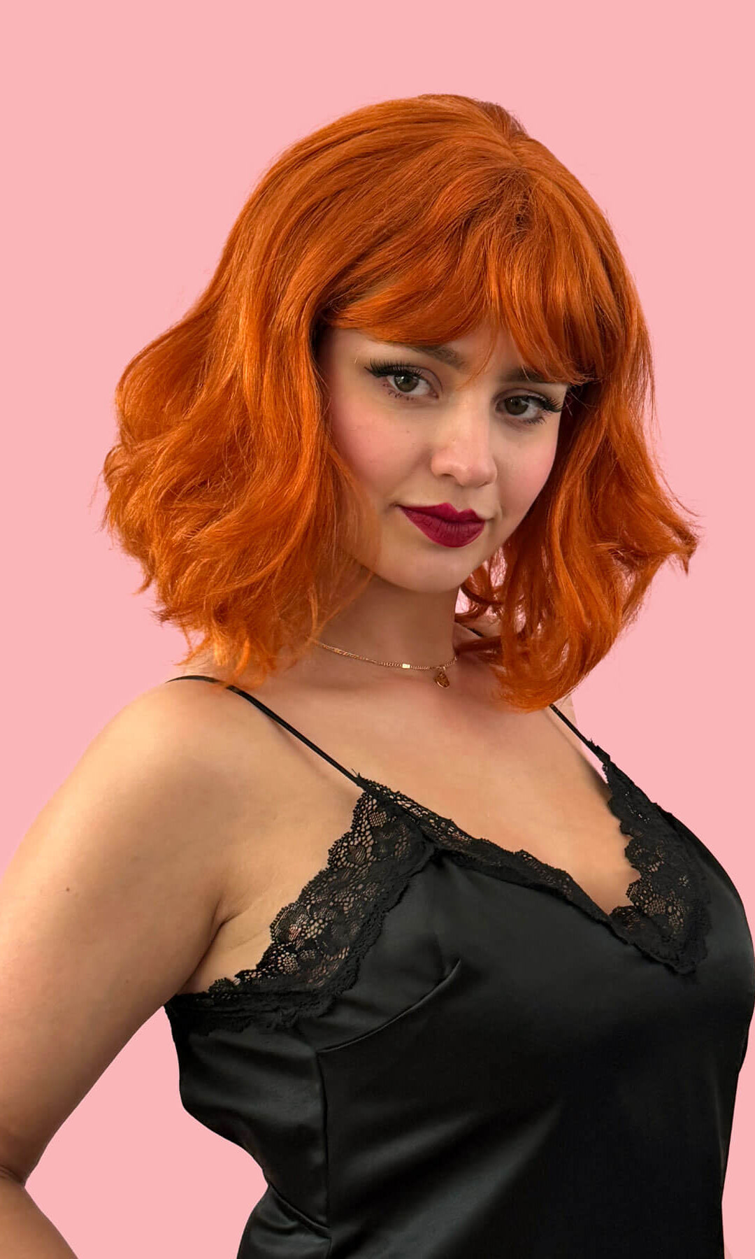 Woman with red hair wearing a black lace top against a pink background. Ginger long wavy bob (lob) wig with swept fringe: Rusty