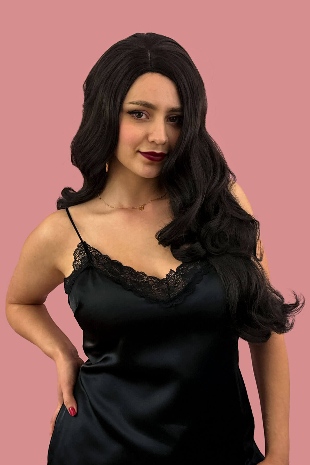Woman wearing a black satin dress with lace straps against a pink background. Long, brown wig, with wonderful loose waves and curls and a sweeping fringe: Leonie