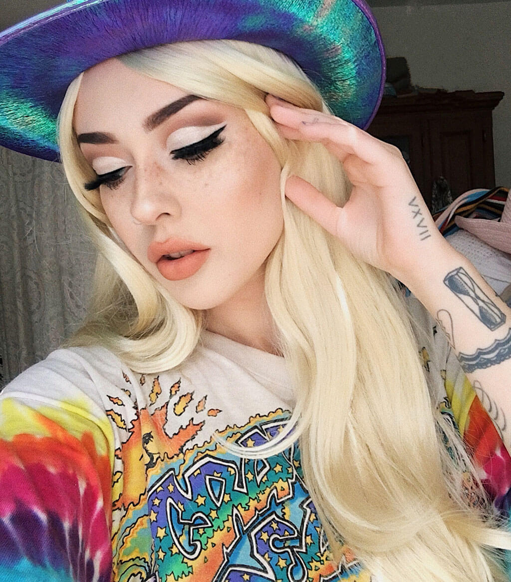 Person with colorful hat and clothing, posing with hand near face. Long straight blonde wig with no fringe: Juste