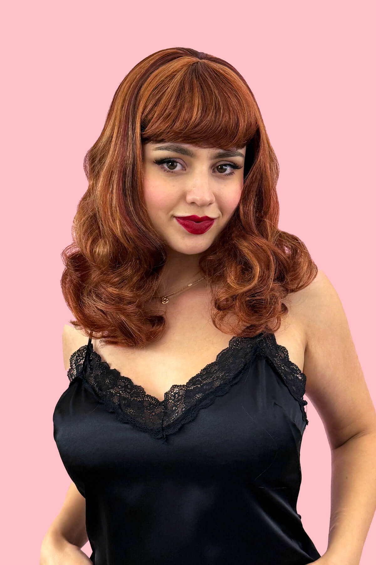 Woman with a vintage-style wig and black lace top against a pink background. Auburn pinup style wig, finger waved with short fringe, 1950s style: Heidi