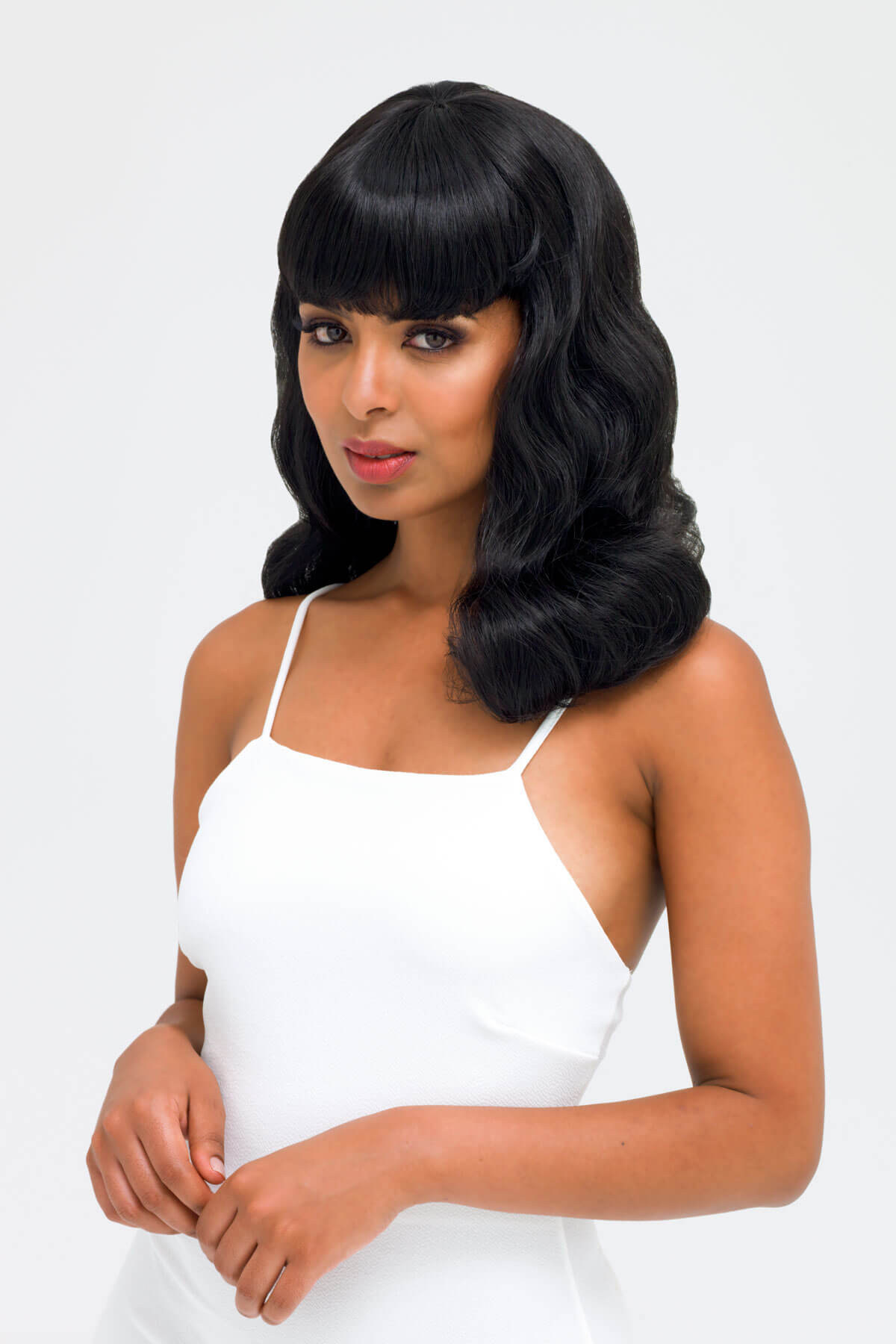 Woman with styled black hair wearing a white dress against a light gray background. Black pinup style wig, curled with short fringe, 1950s style: Bettie