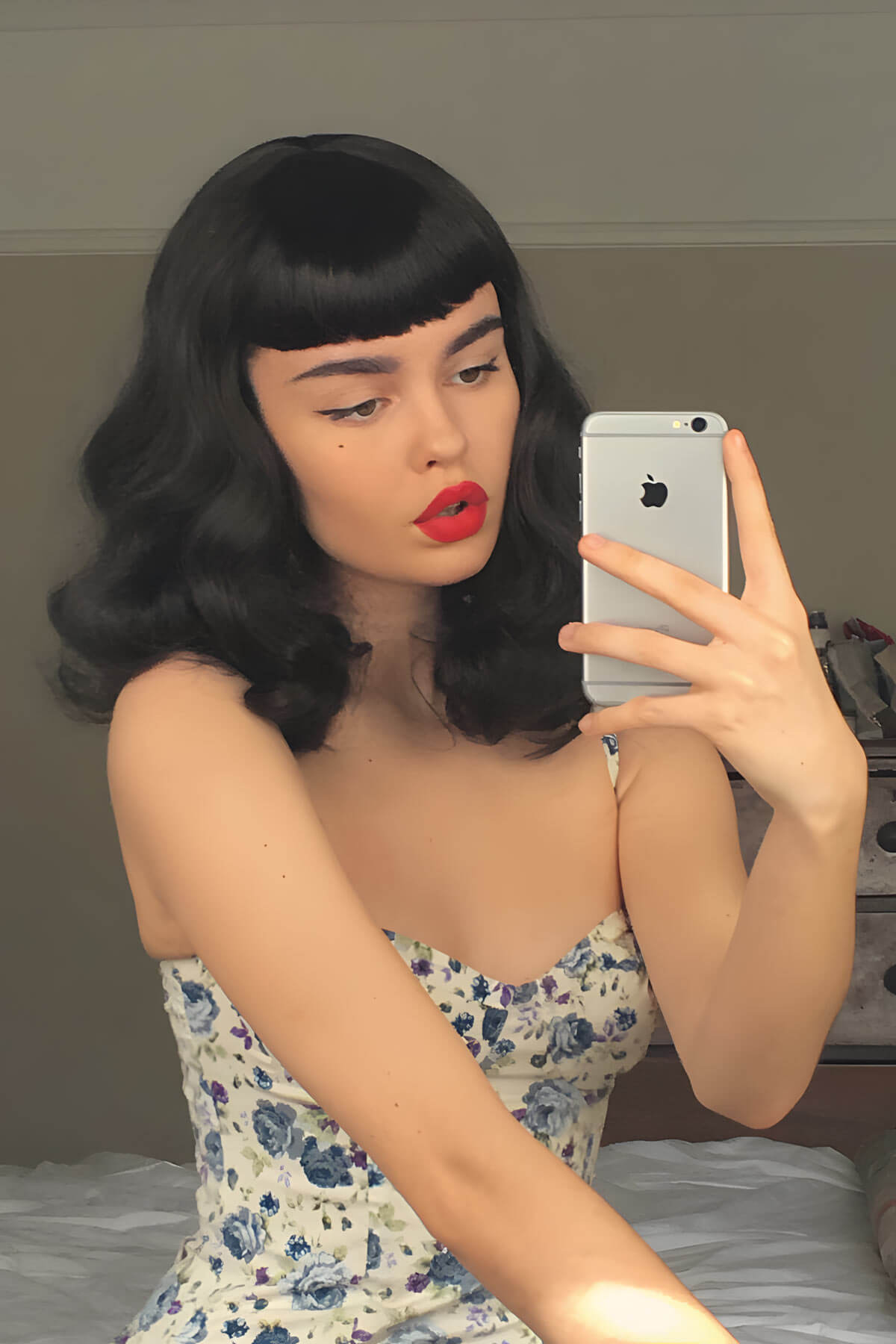 Woman with black hair and red lipstick taking a mirror selfie with a floral dress. Black pinup style wig, curled with short fringe, 1950s style: Bettie