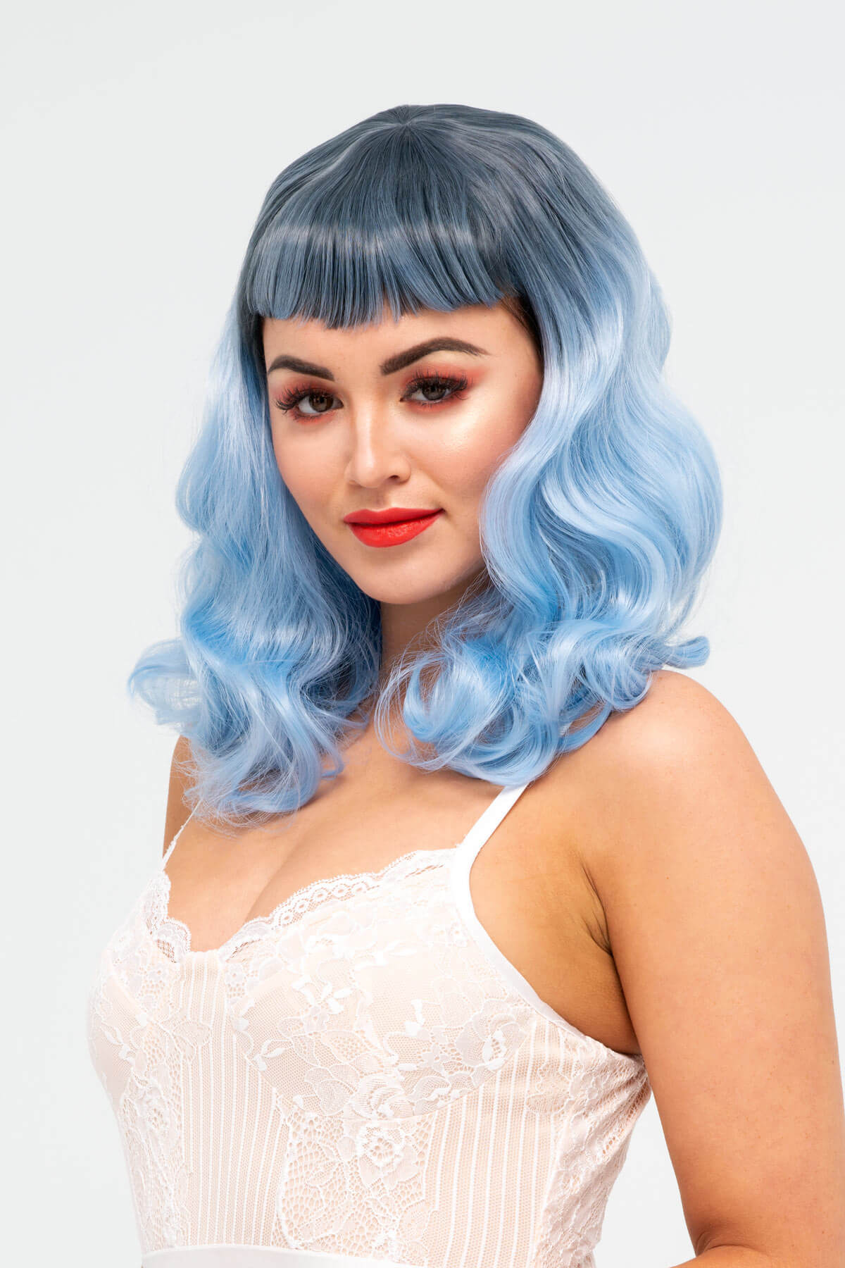 Woman with blue hair styled in waves wearing a white lace top against a light gray background. Black and blue pinup style wig, curled with short fringe, 1950s style: Luna