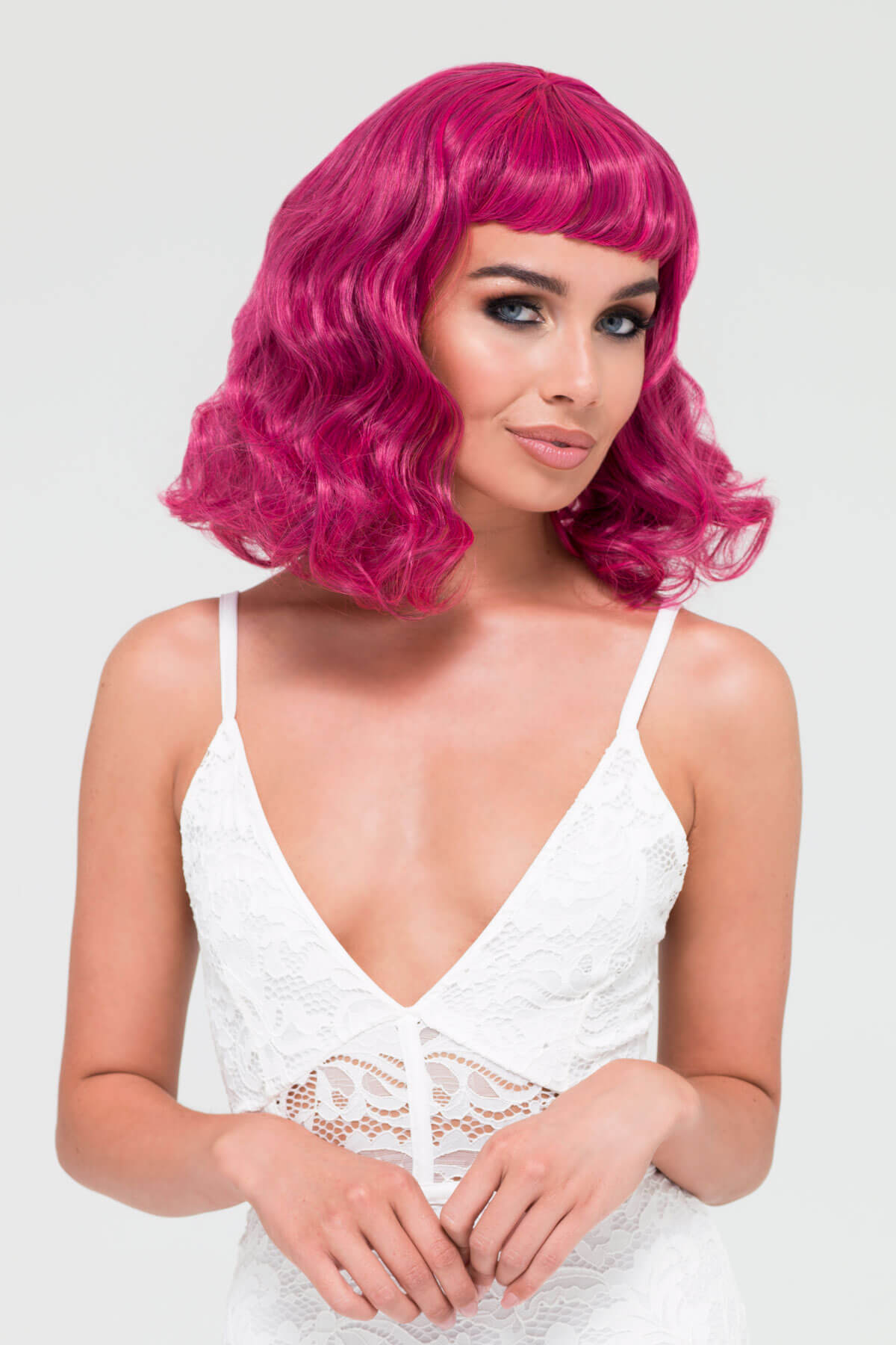 Woman with bright pink wig wearing a white lace top on a light gray background. Black and pink pinup wig, curled with a short fringe, 1950s style: Billie