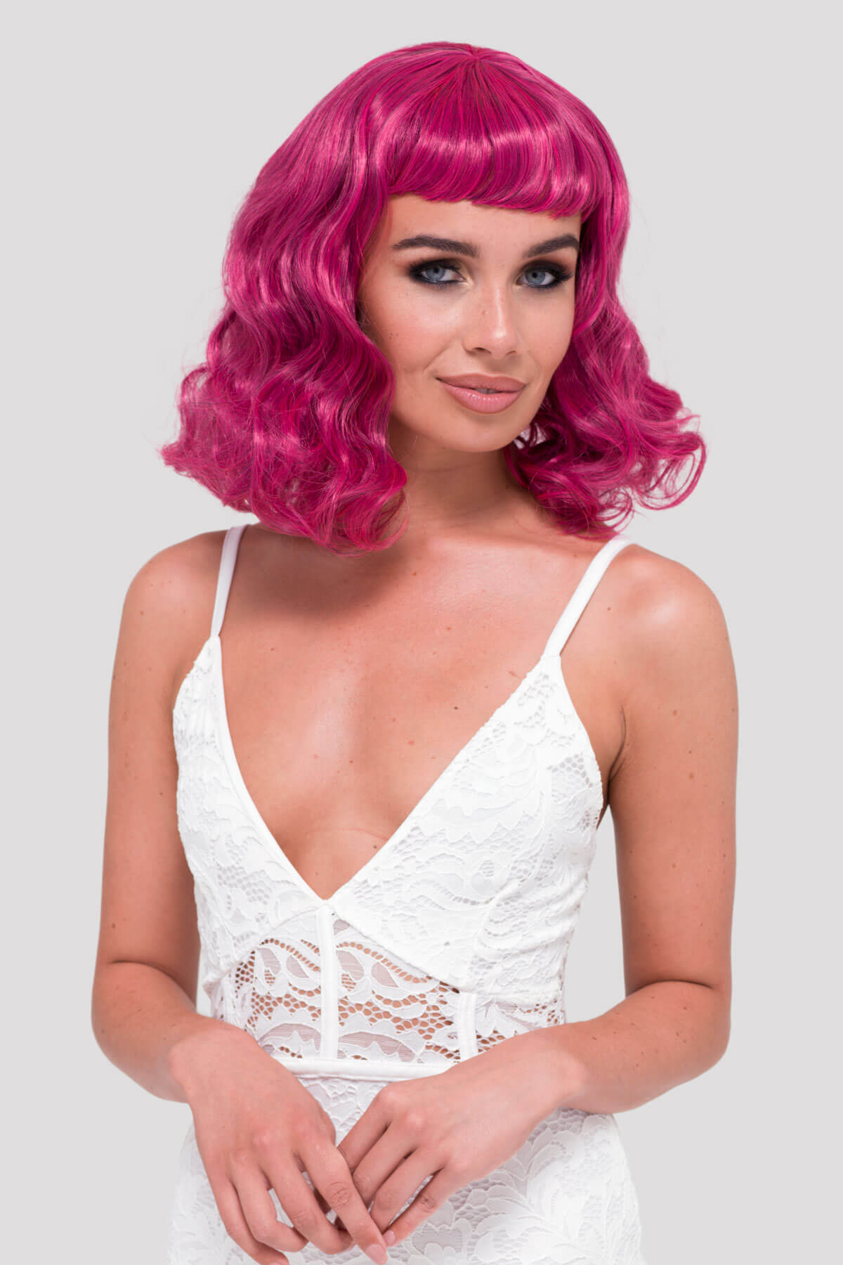 Woman with a bright pink wig wearing a white lace top on a light gray background. Black and pink pinup wig, curled with a short fringe, 1950s style: Billie