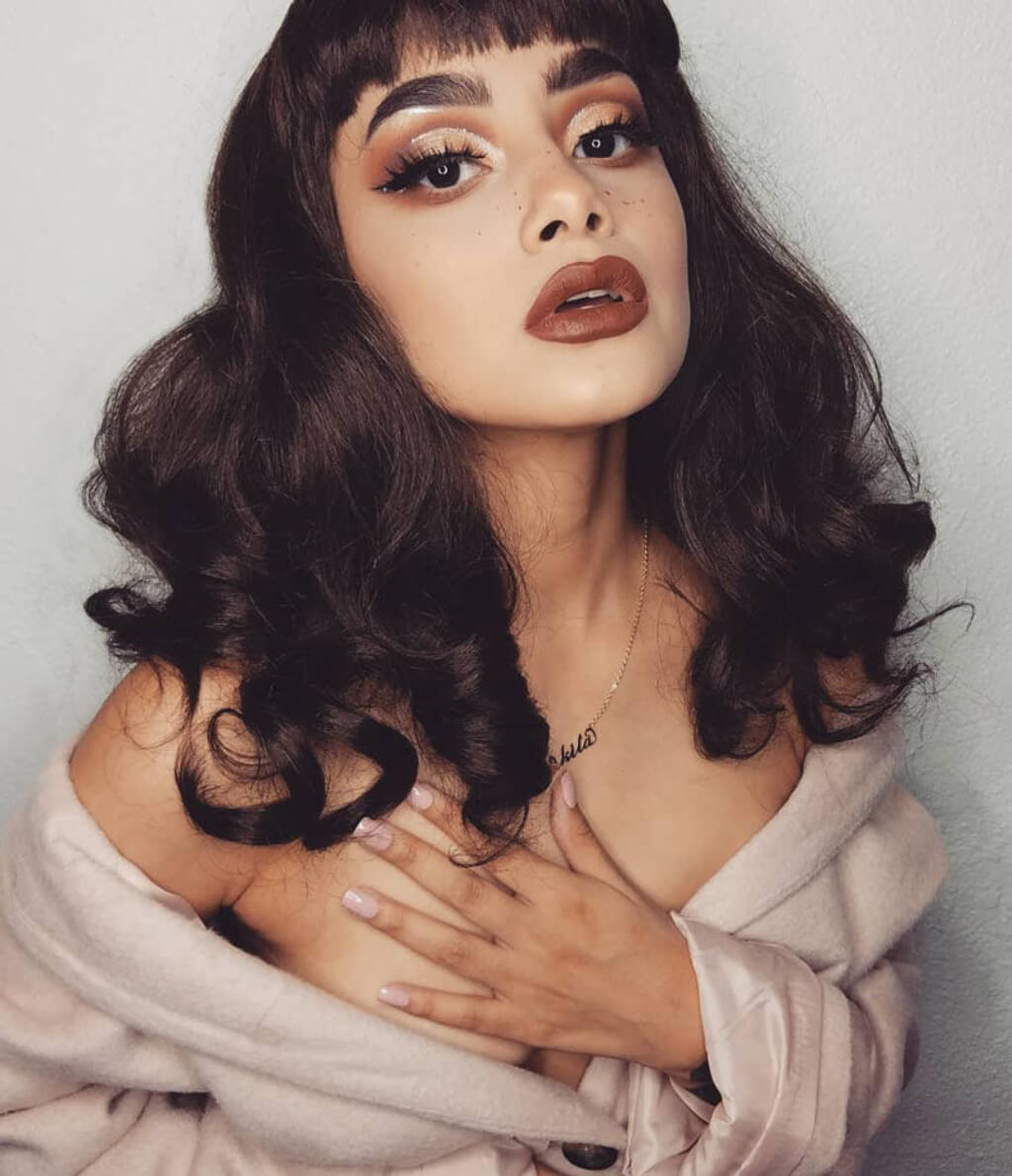 A woman with long, dark brown wavy hair and bangs, wearing a light pink satin robe. Dark brown pinup style wig, finger waved with short fringe, 1950s style: Becca