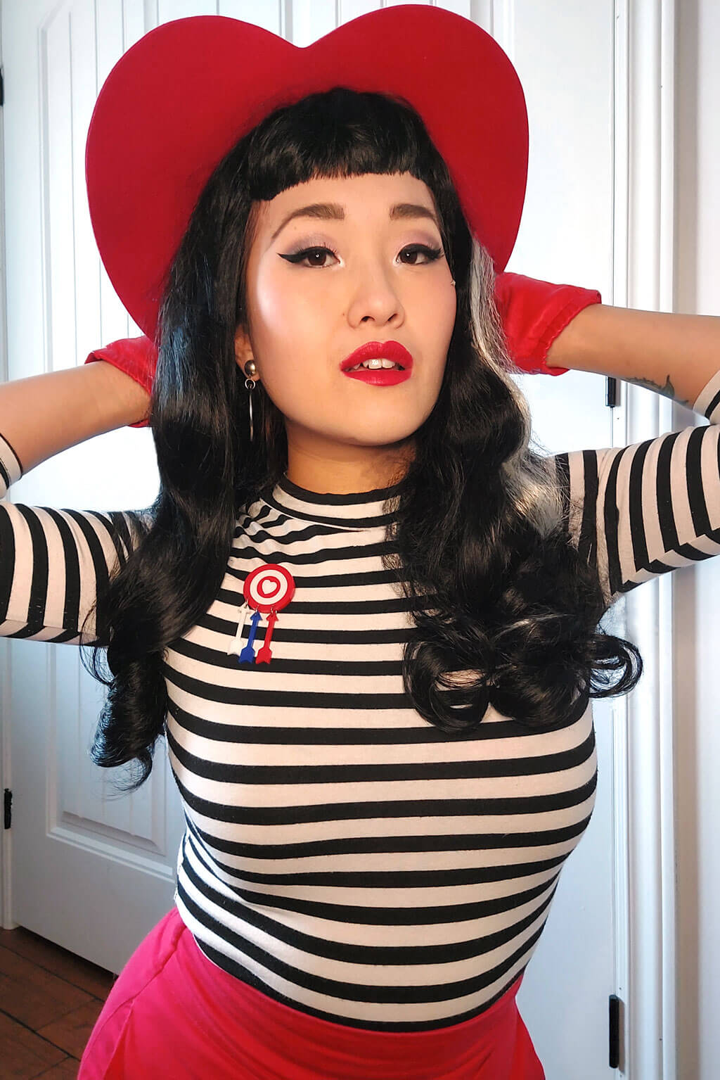 Person wearing a red heart-shaped hat, striped shirt, and pink skirt indoors. Long black 1950s pin up style wig with platinum streak and retro fringe: Michelle