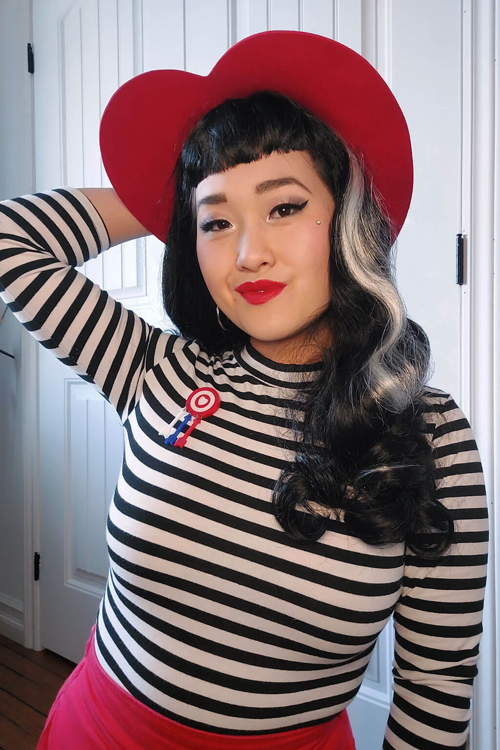 Person wearing a black and white striped shirt with a red heart-shaped hat and red skirt. Long black 1950s pin up style wig with platinum streak and retro fringe: Michelle