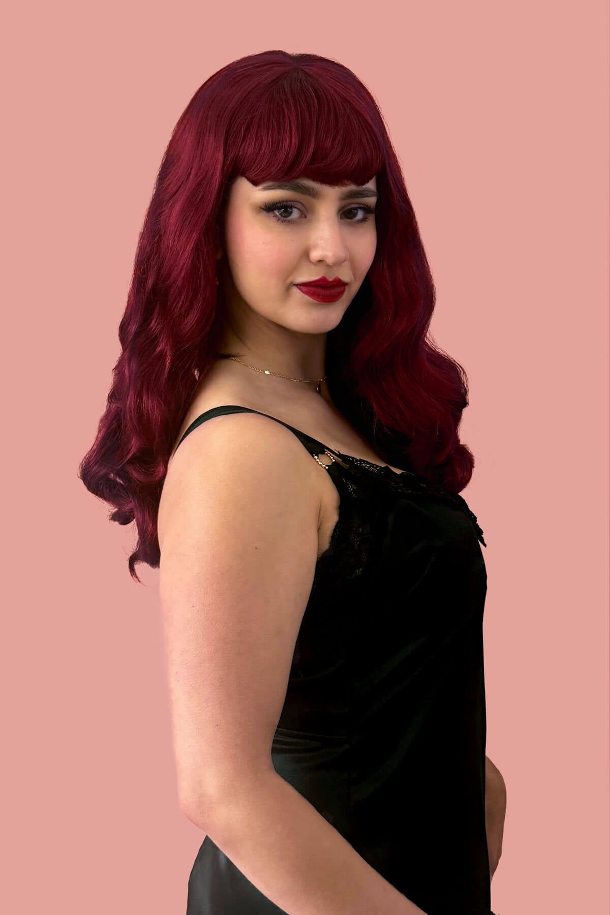 Woman with vibrant red hair and red lipstick wearing a black dress against a pink background. Long cherry red pin-up wig, gently wavy with short fringe: Maeve