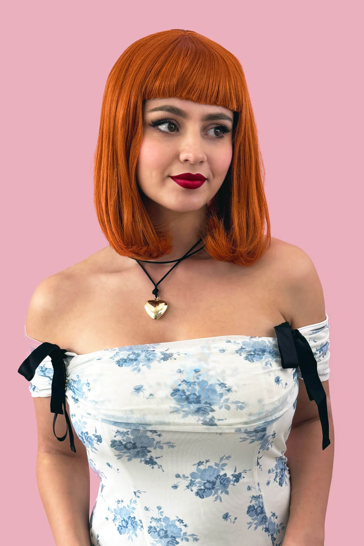 Woman with red hair wearing a white floral dress with black straps against a pink background. Ginger long bob wig with a short retro style fringe: Bonnie