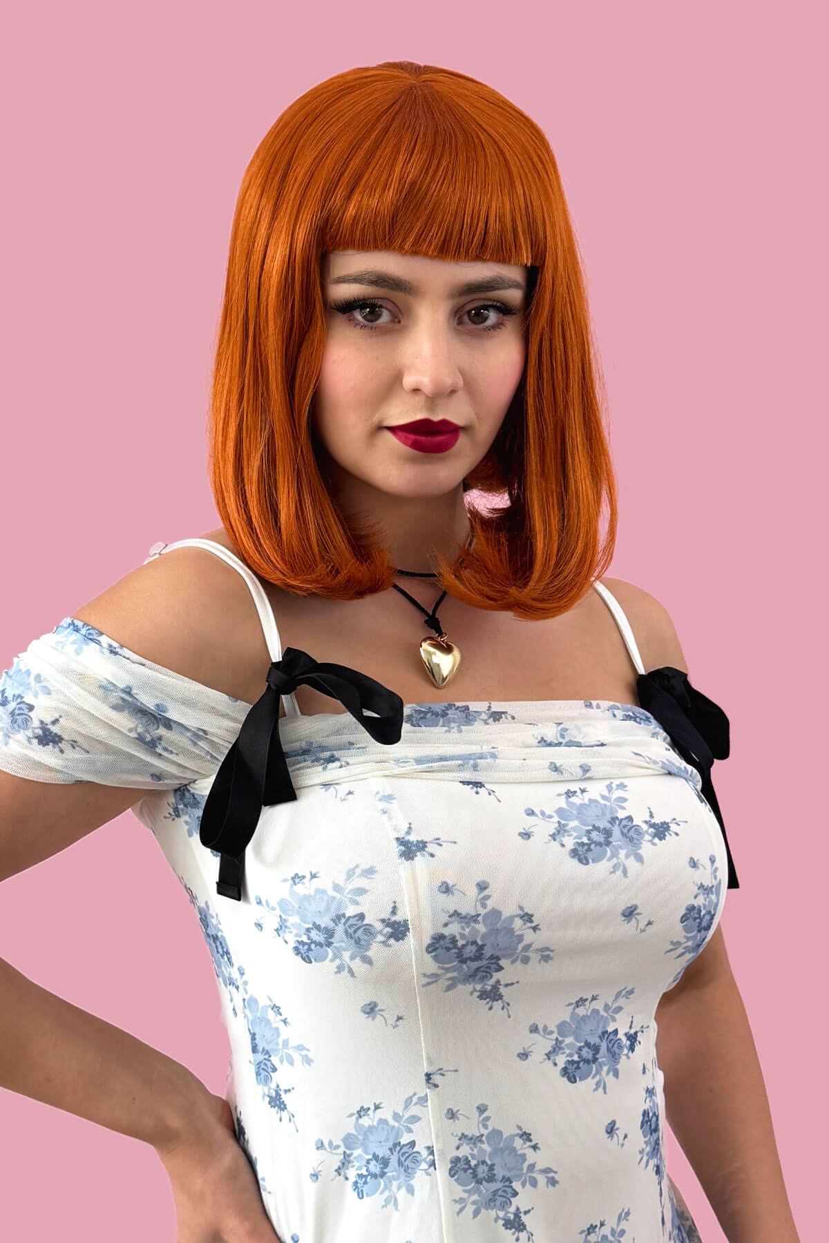 Woman with orange wig and floral dress against a pink background. Ginger long bob wig with a short retro style fringe: Bonnie