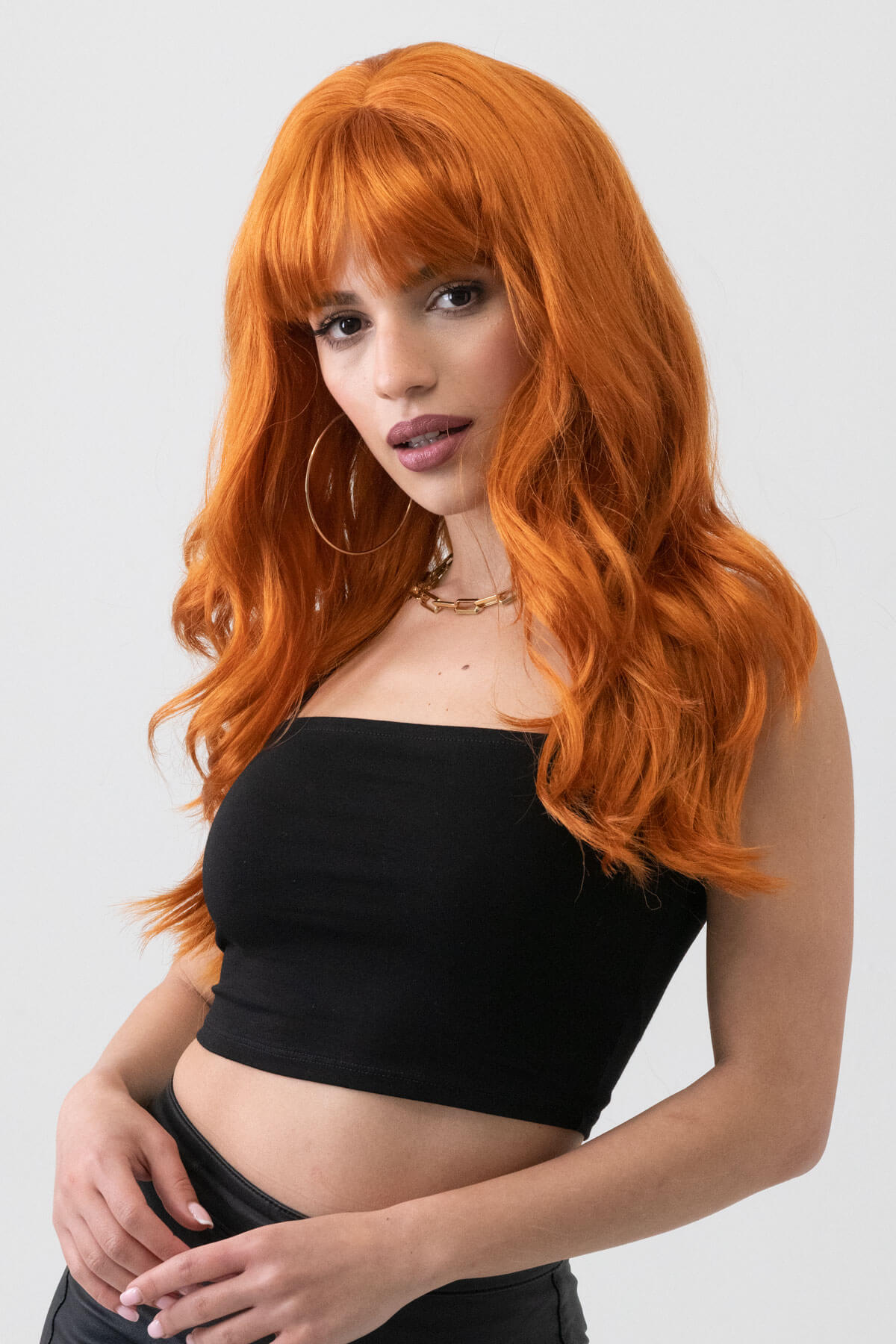 Woman with long orange hair wearing a black top against a light gray background. Long ginger wig with cascading waves: Davina