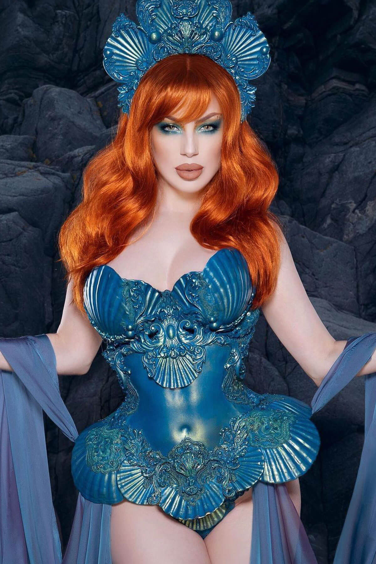 Woman in a blue costume with intricate designs against a dark background. Long ginger wig with cascading waves: Davina