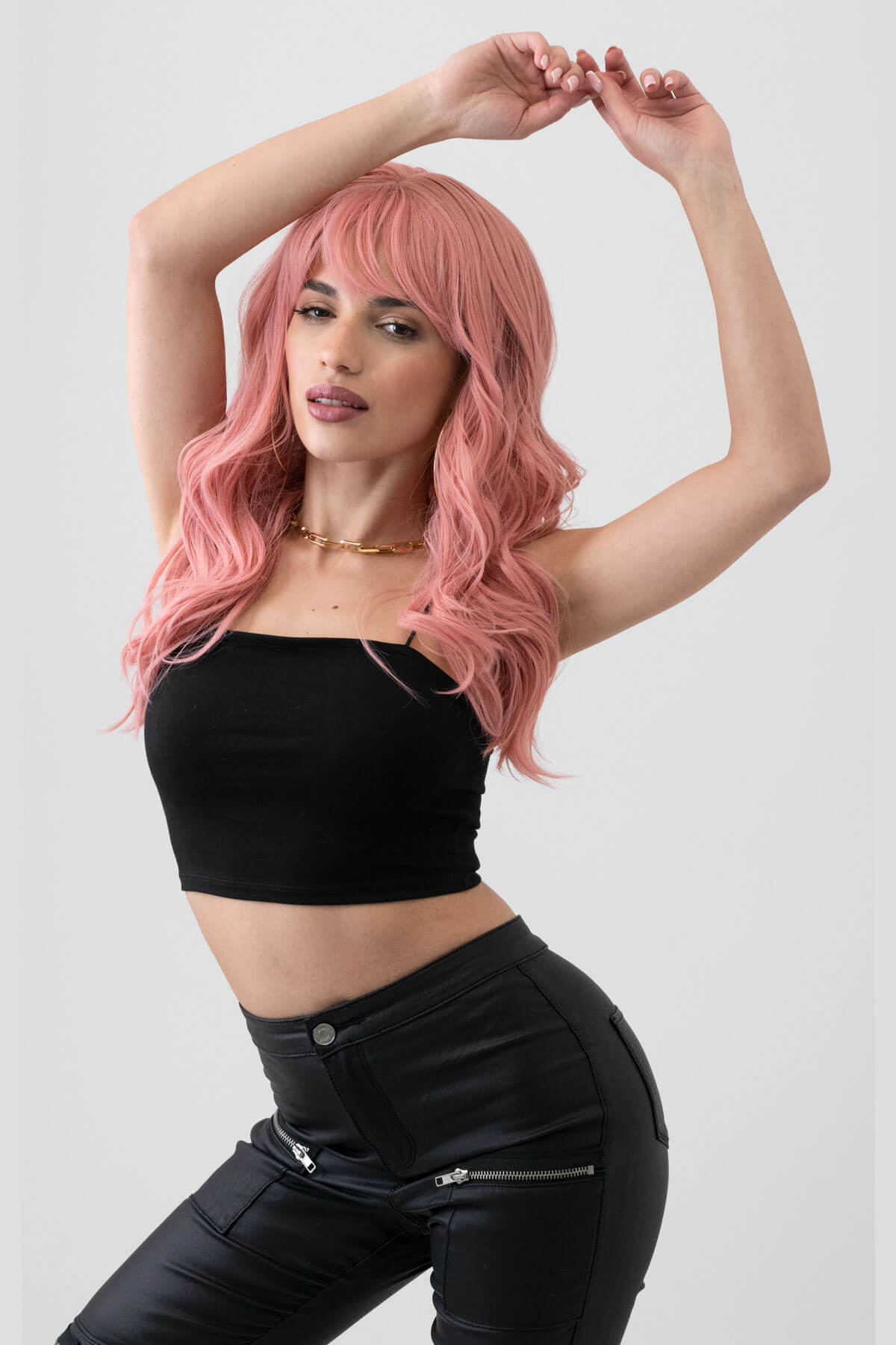 Woman with pink hair wearing a black crop top and black pants on a white background. Dusty pastel pink wig with long cascading waves: Rosalie