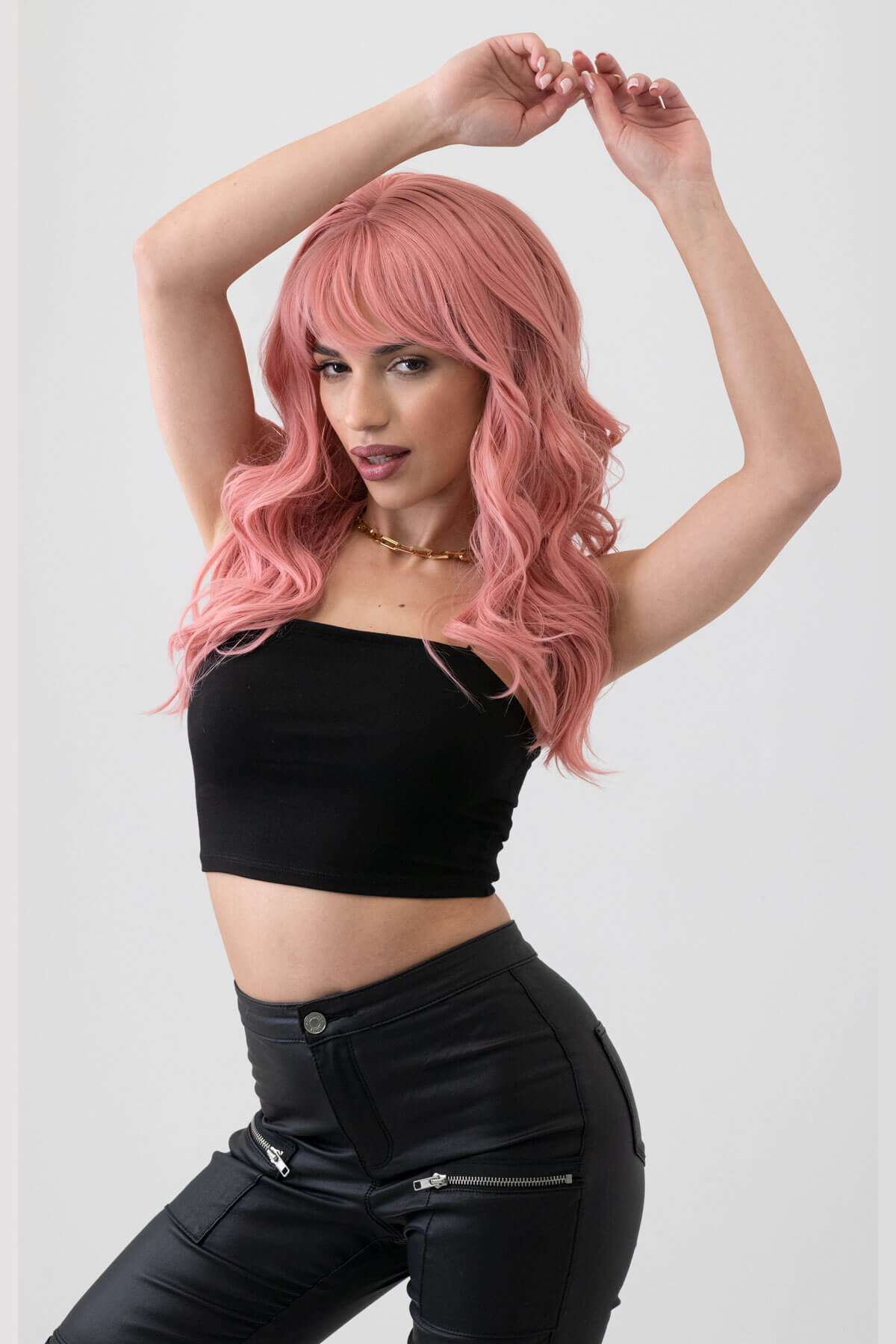 Woman with pink hair wearing a black top and pants on a white background. Dusty pastel pink wig with long cascading waves: Rosalie