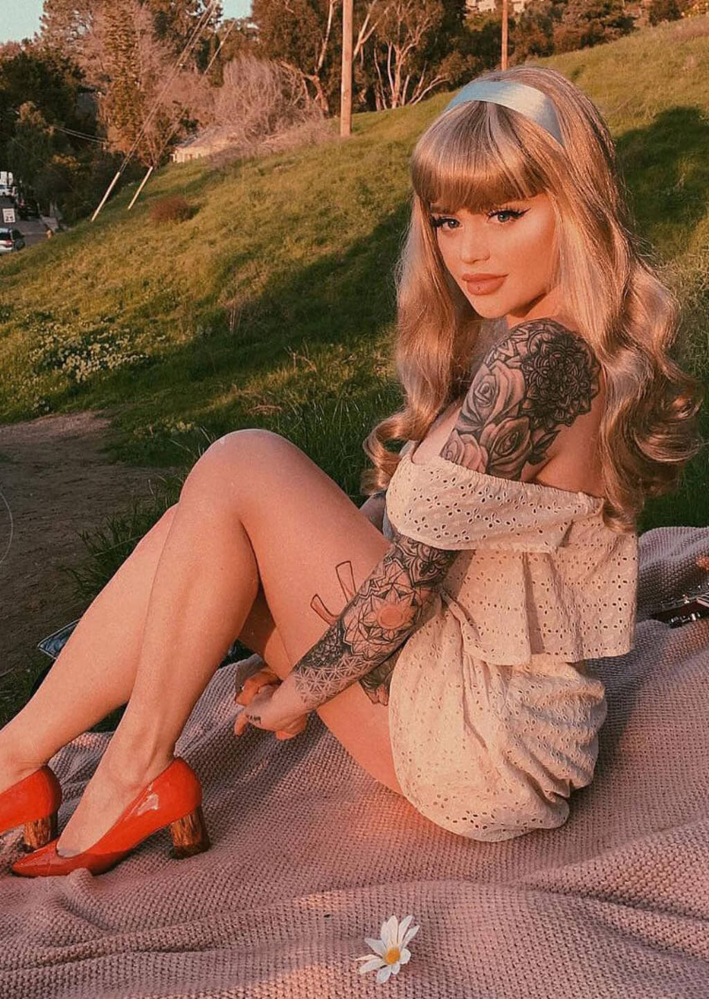Woman with tattoos sitting outdoors on a blanket. Long ash blonde wig with gentle waves: Harriet