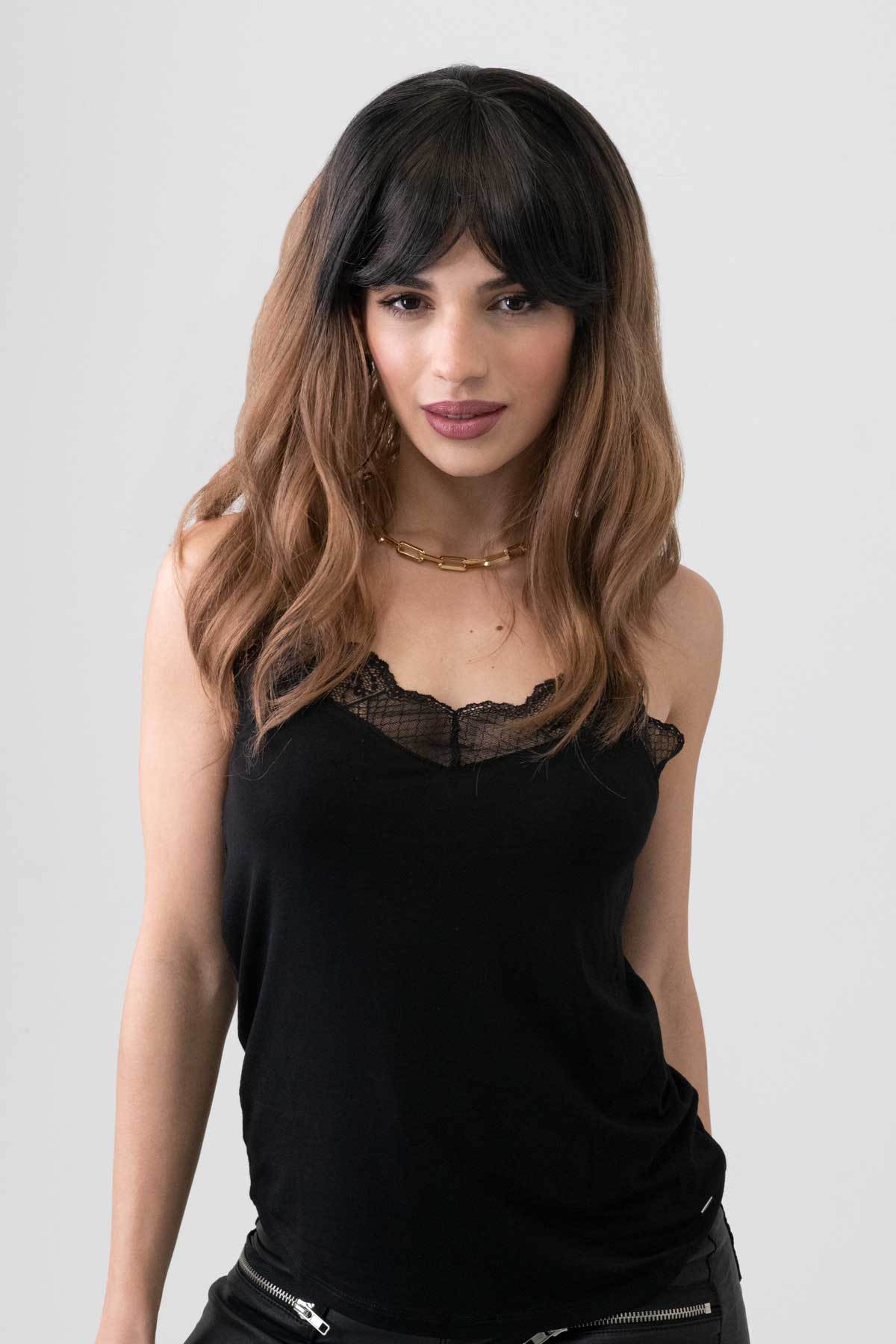 Woman wearing a black sleeveless top with lace details on a plain background. Long wavy bob wig (lob), dark brown to light brown ombre: Rowena