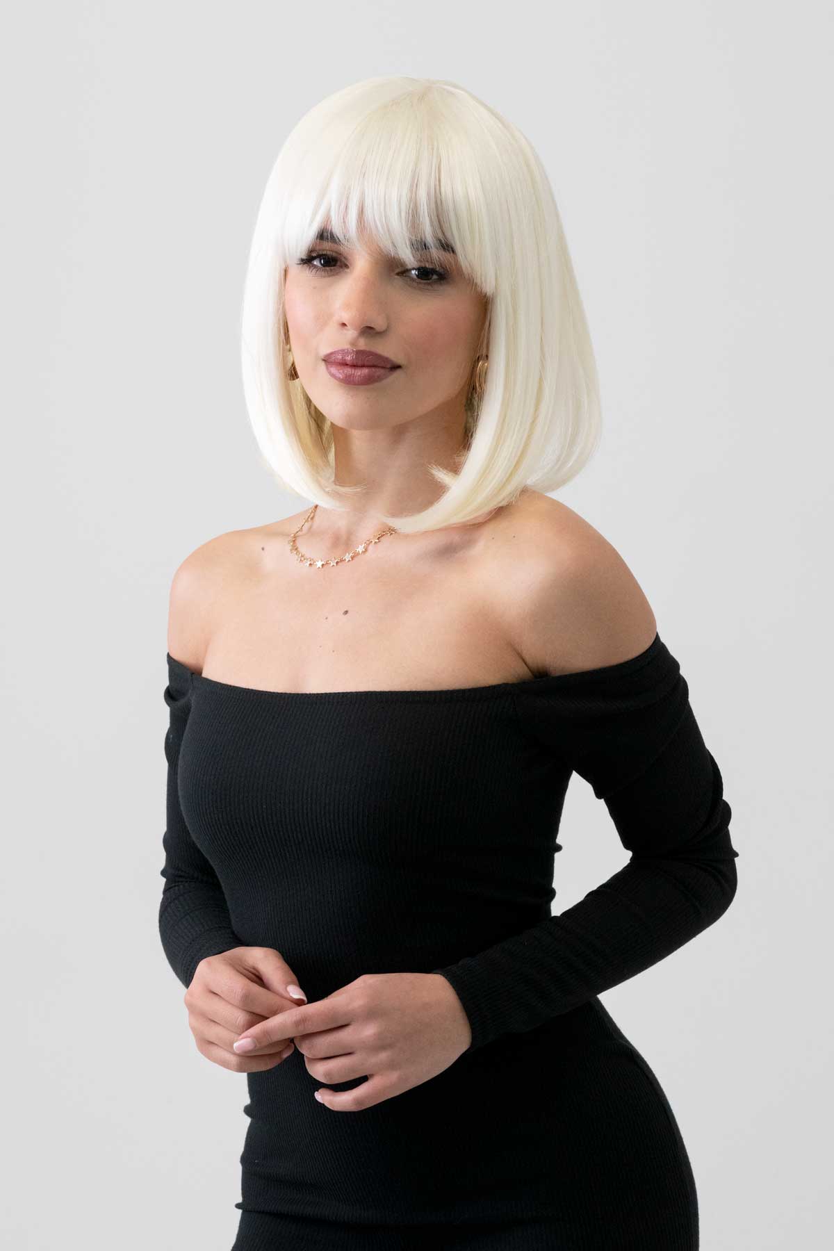 Woman with platinum blonde hair wearing a black off-shoulder dress with a plain background. Platinum blonde bob wig, shoulder length: Neve