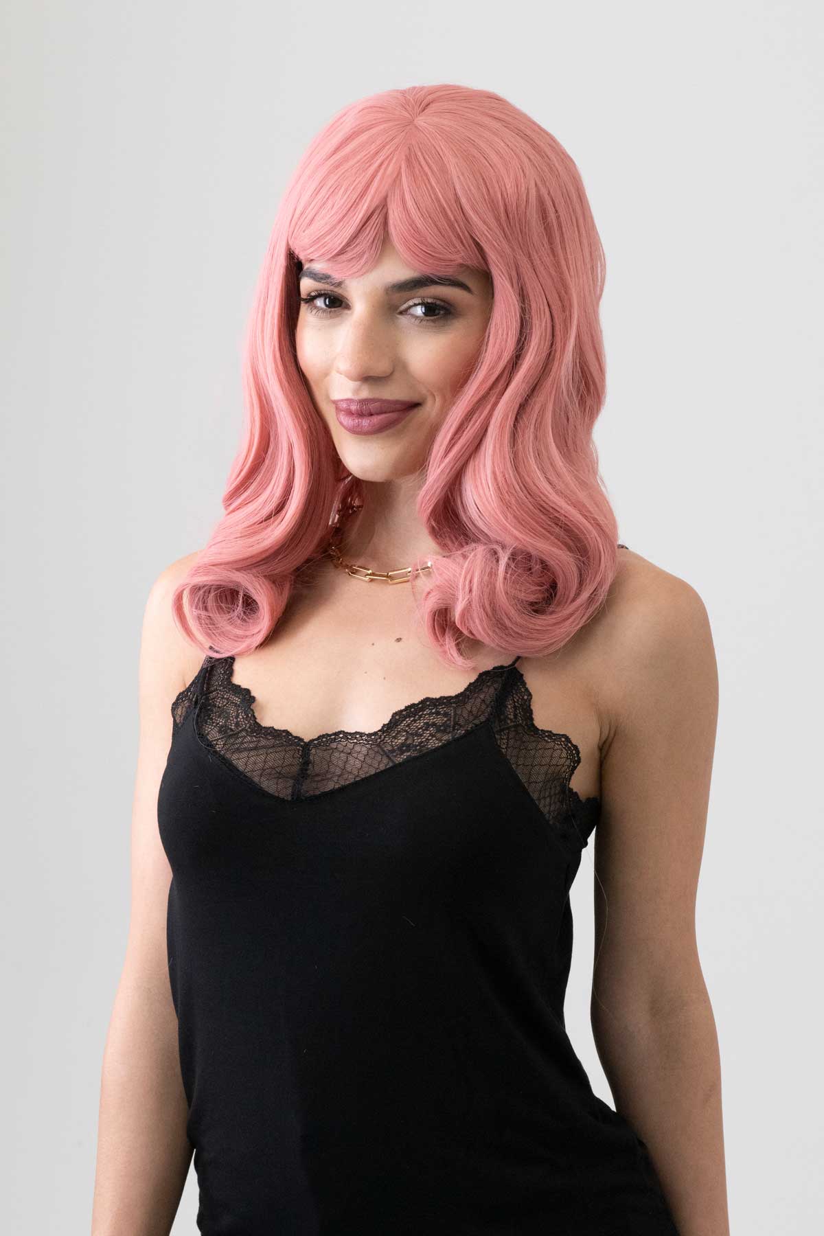 Woman with pink wig wearing a black lace top against a gray background. Dusty Pink 50s vintage style wig with short fringe: Paloma