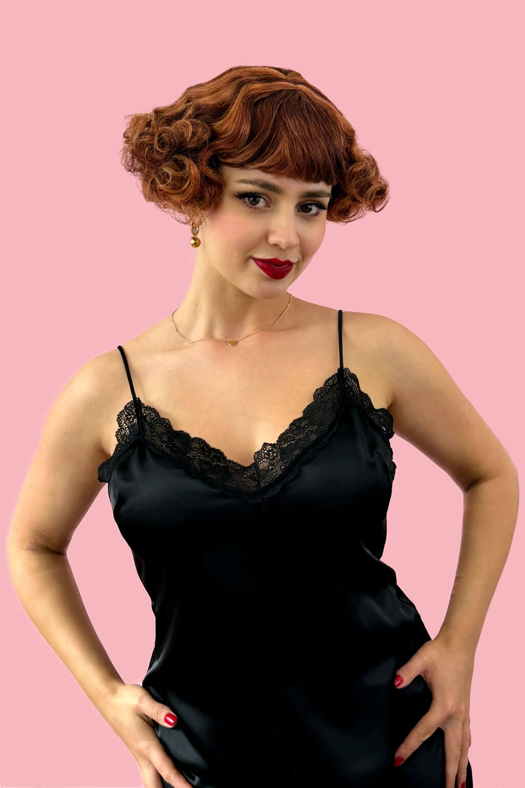 Woman wearing a black satin dress against a pink background. A short red, auburn and blonde mix vintage style wig with waves: Solana
