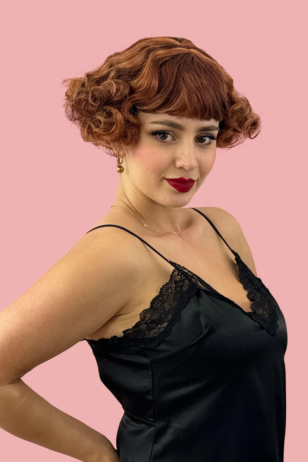 Woman wearing a black satin dressagainst a pink background. A short red, auburn and blonde mix vintage style wig with waves: Solana