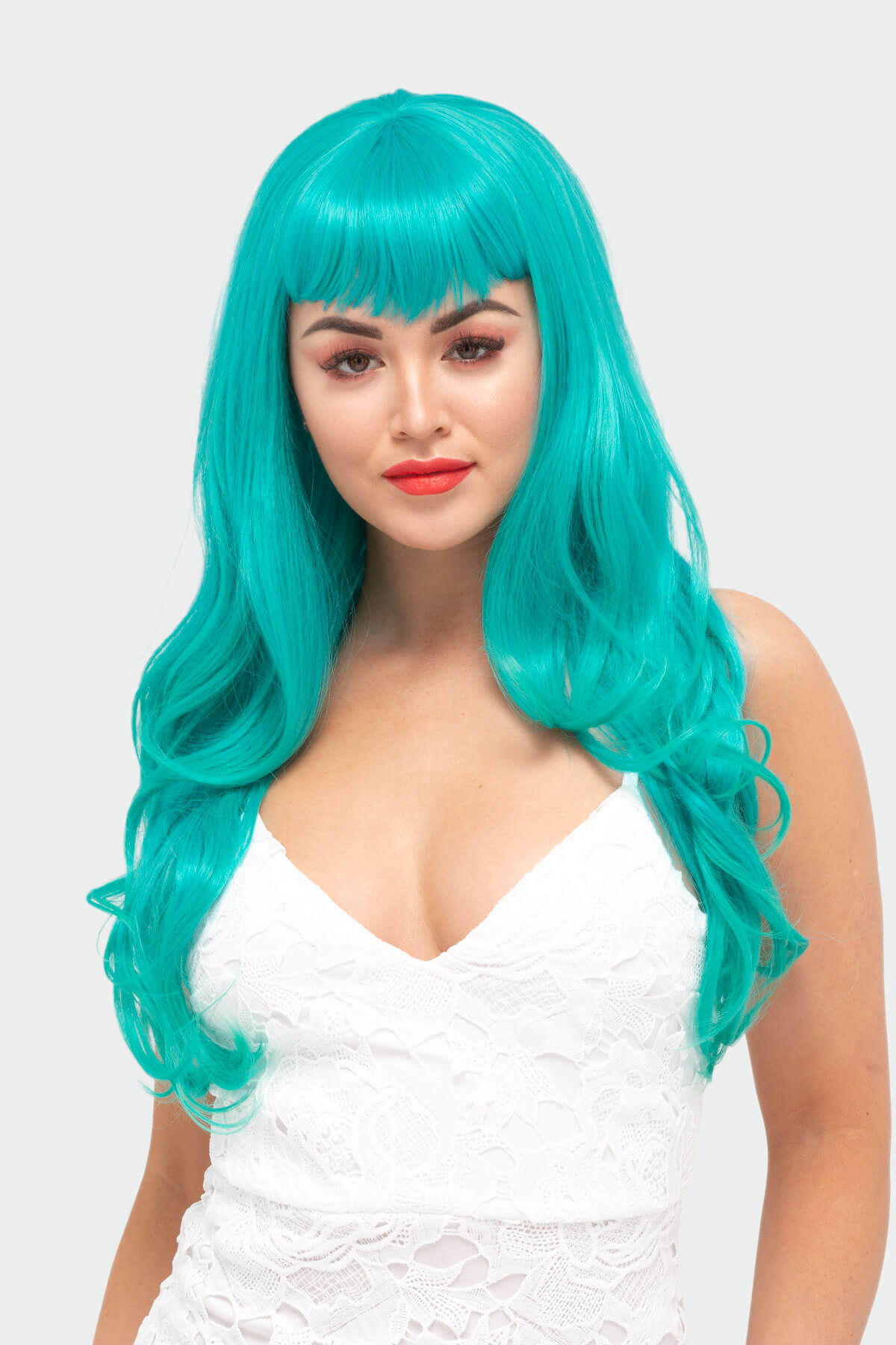 Woman with teal wig and white dress on a light gray background. Long aqua green wig with gentle waves and vintage-style straight fringe: Natalie
