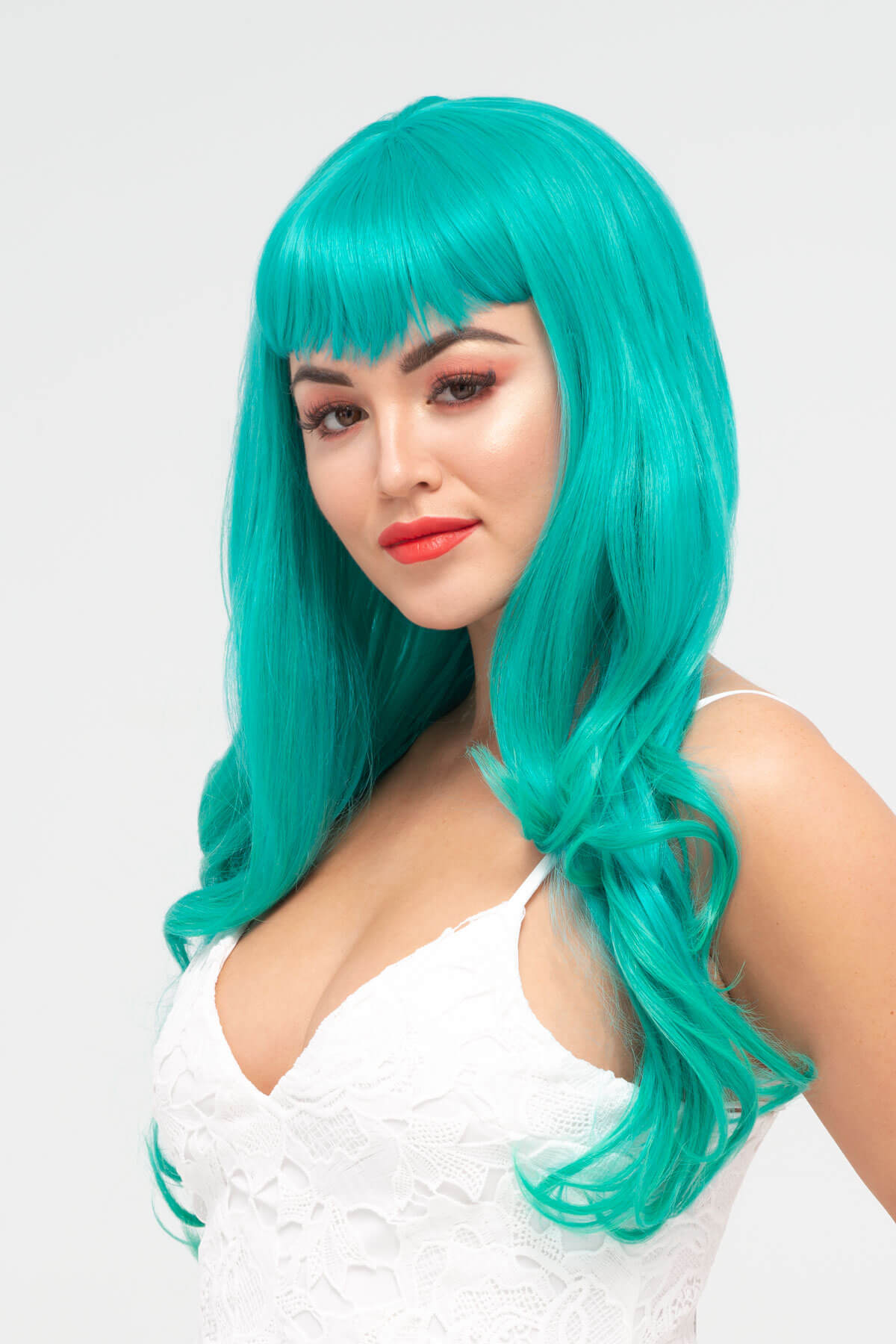 Woman with teal wig and white top on a light gray background. Long aqua green wig with gentle waves and vintage-style straight fringe: Natalie