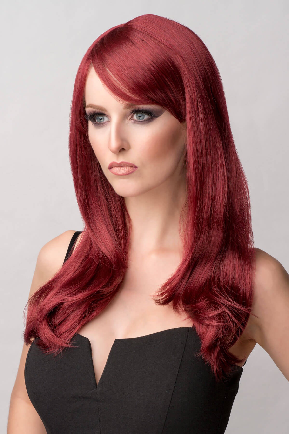 Woman with long red hair wearing a black dress against a gray background. Long red wig in razor cut, face frame style: Megan