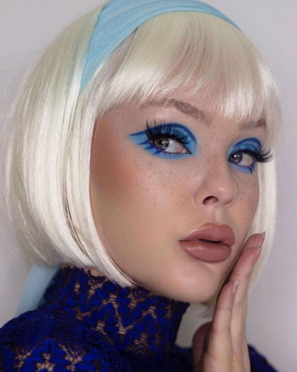 Person with blonde bob haircut and blue headband wearing blue makeup against a white background. Platinum blonde bob wig: Carol-Anne