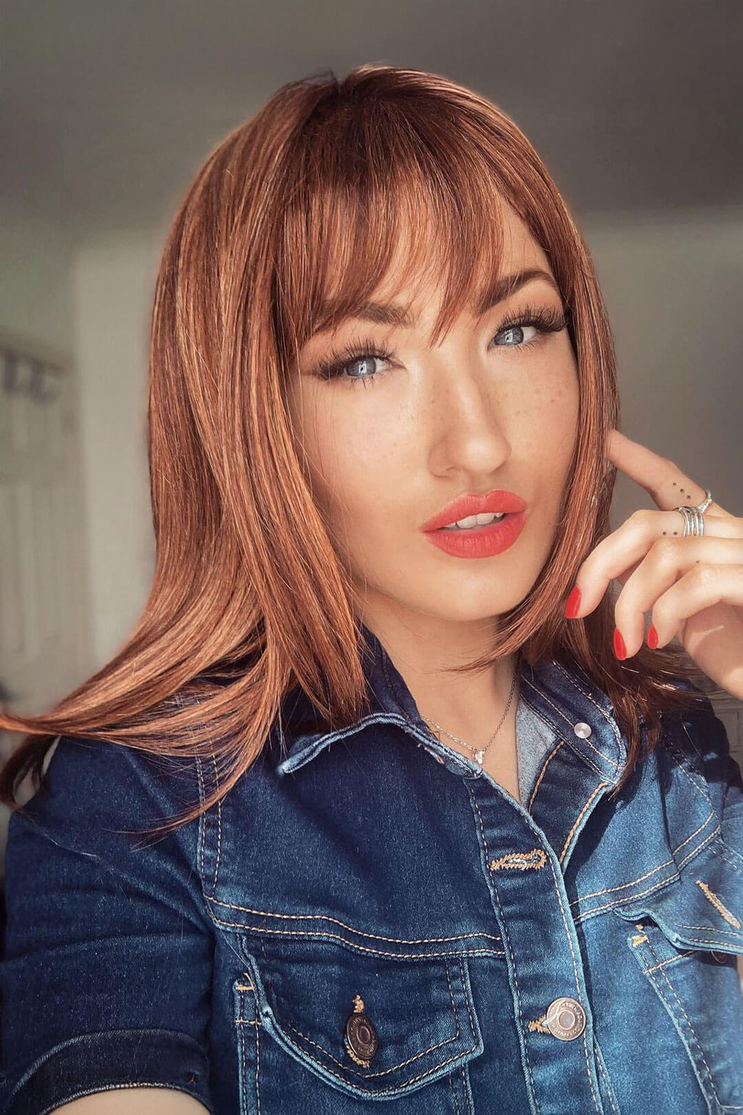 Woman with long auburn hair and denim jacket indoors. Gingery auburn wig with blonde highlights face frame style: Geri