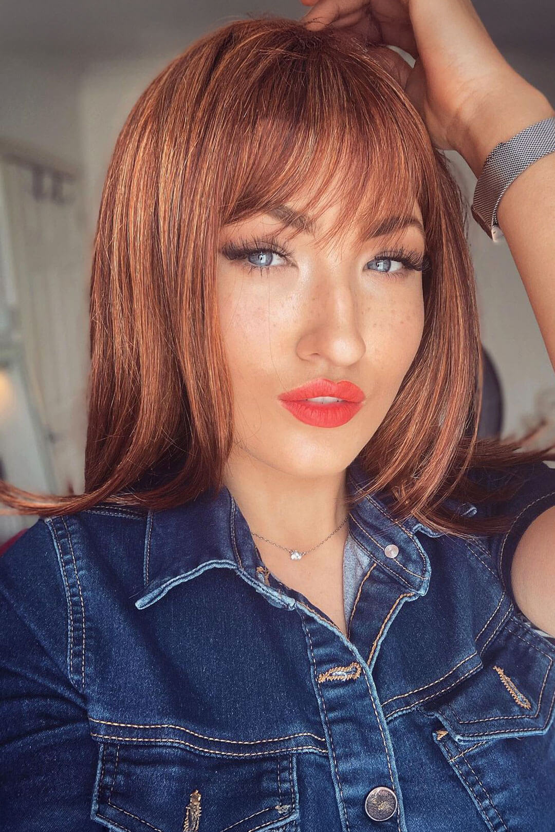 Woman with styled auburn hair and denim jacket in a blurred indoor setting. Gingery auburn wig with blonde highlights face frame style: Geri