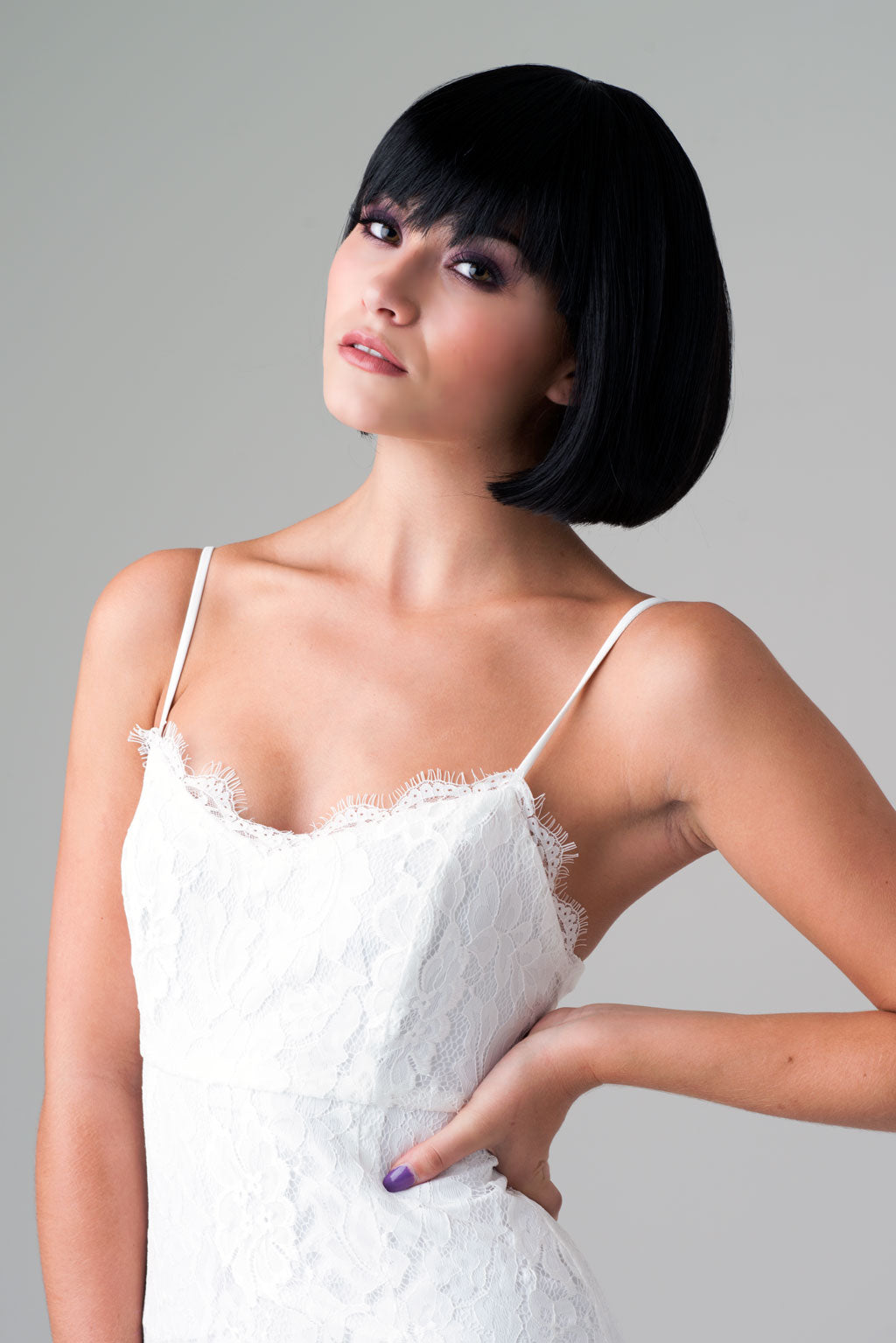 Woman wearing a white lace dress with a plain background. Short black bob wig: Josie
