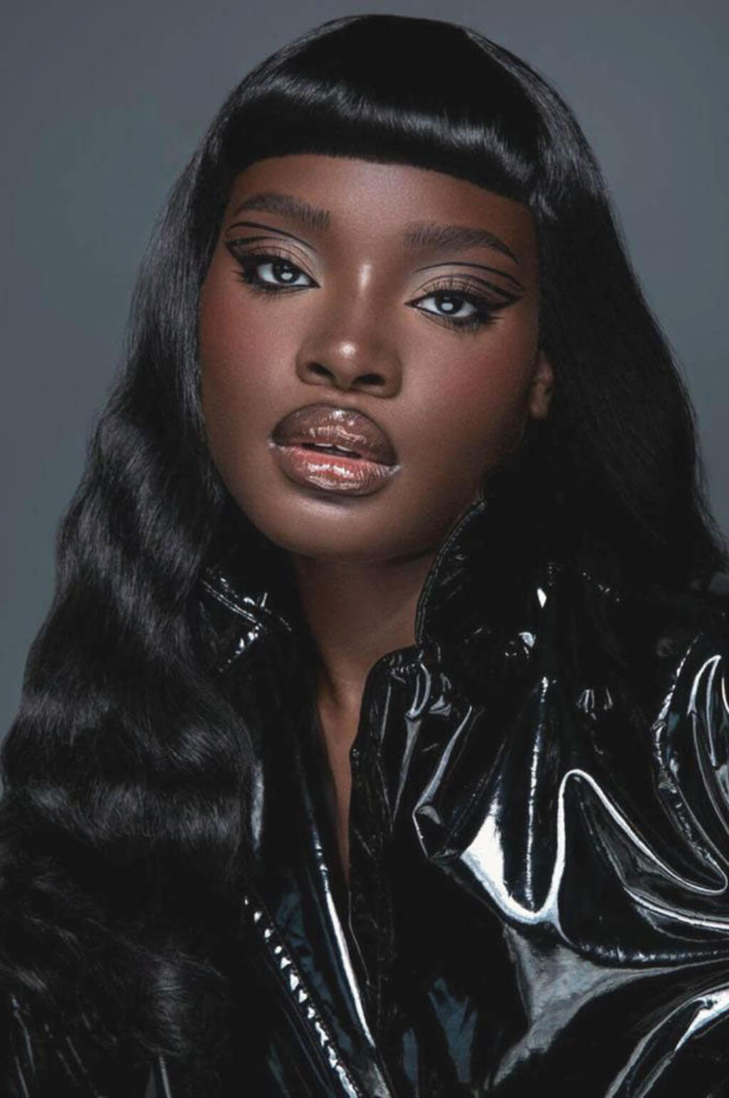 Woman wearing a black glossy outfit against a grey background. Black 1950s pinup style wig, curled with short fringe: Milena