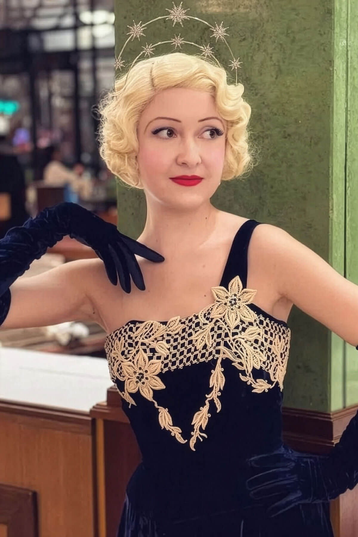 Woman in a vintage-style dress with lace detailing and black gloves, standing indoors. Blonde pinup wig, lace front, vintage style: Odette champagne blonde 613