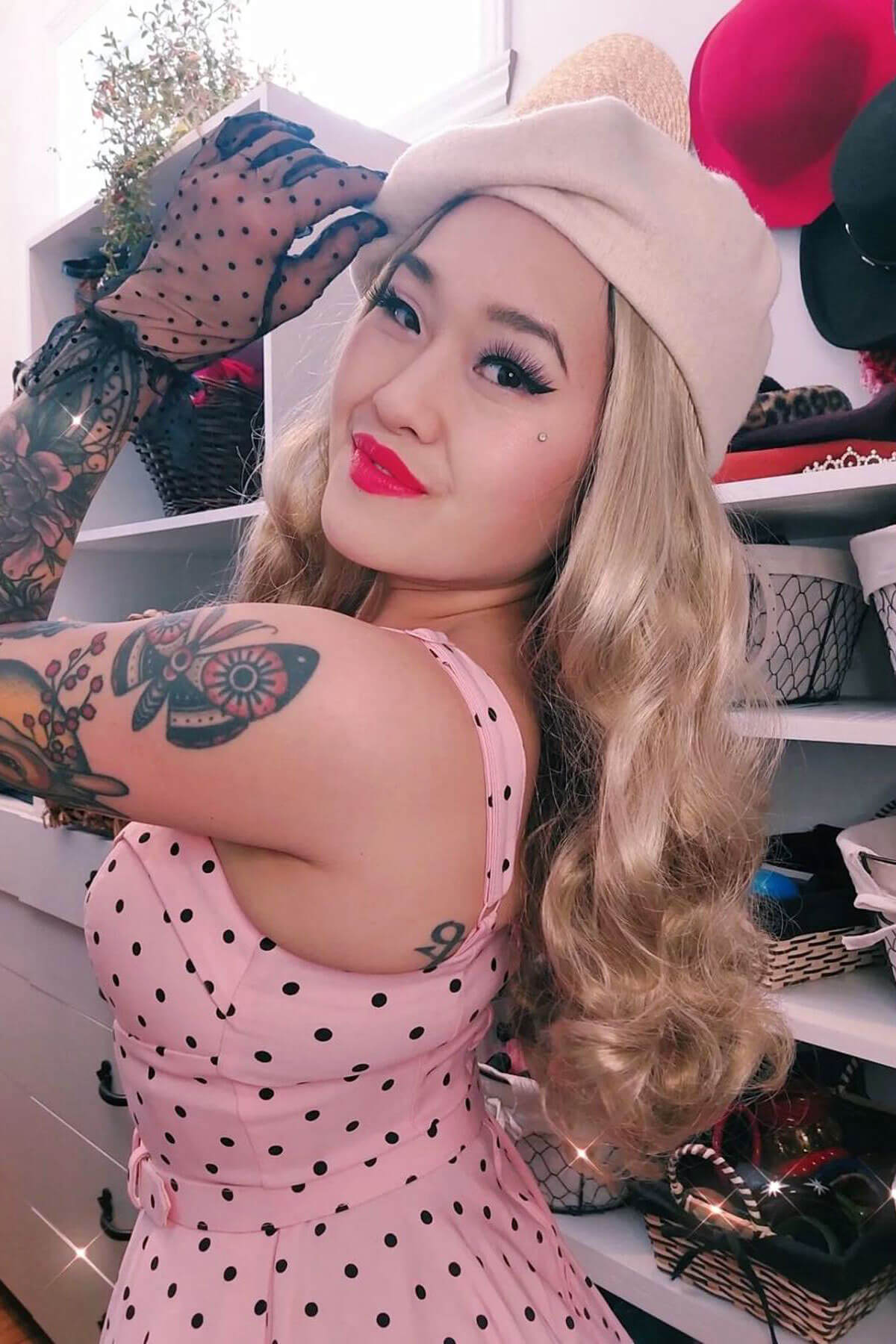Woman in a pink polka dot dress with tattoos and a tattooed arm, wearing a beige beret, in a room with shelves and various items. Blonde wig with waves and a side parting, 1940s style: Nancy