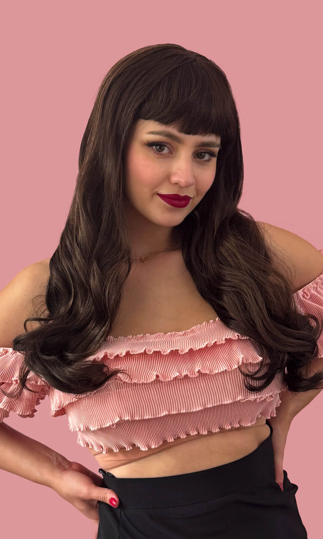 A woman wearing a long chestnut brown wig with blonde highlights and a pinup-style fringe, posing in front of a pink background. Long chestnut brown wig with blonde highlights and pinup-style fringe: Zola