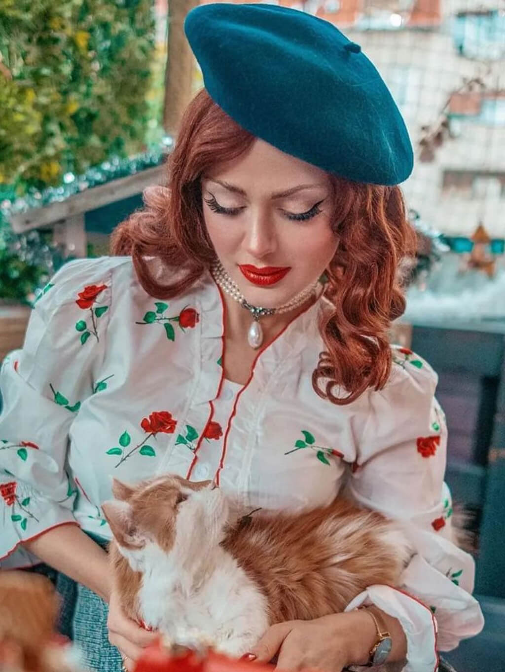A woman in a retro-style outfit with a teal beret and a white floral blouse. The background is a softly blurred outdoor setting. Copper red 40s style wig, wavy: Banbury copper red 40s style wig