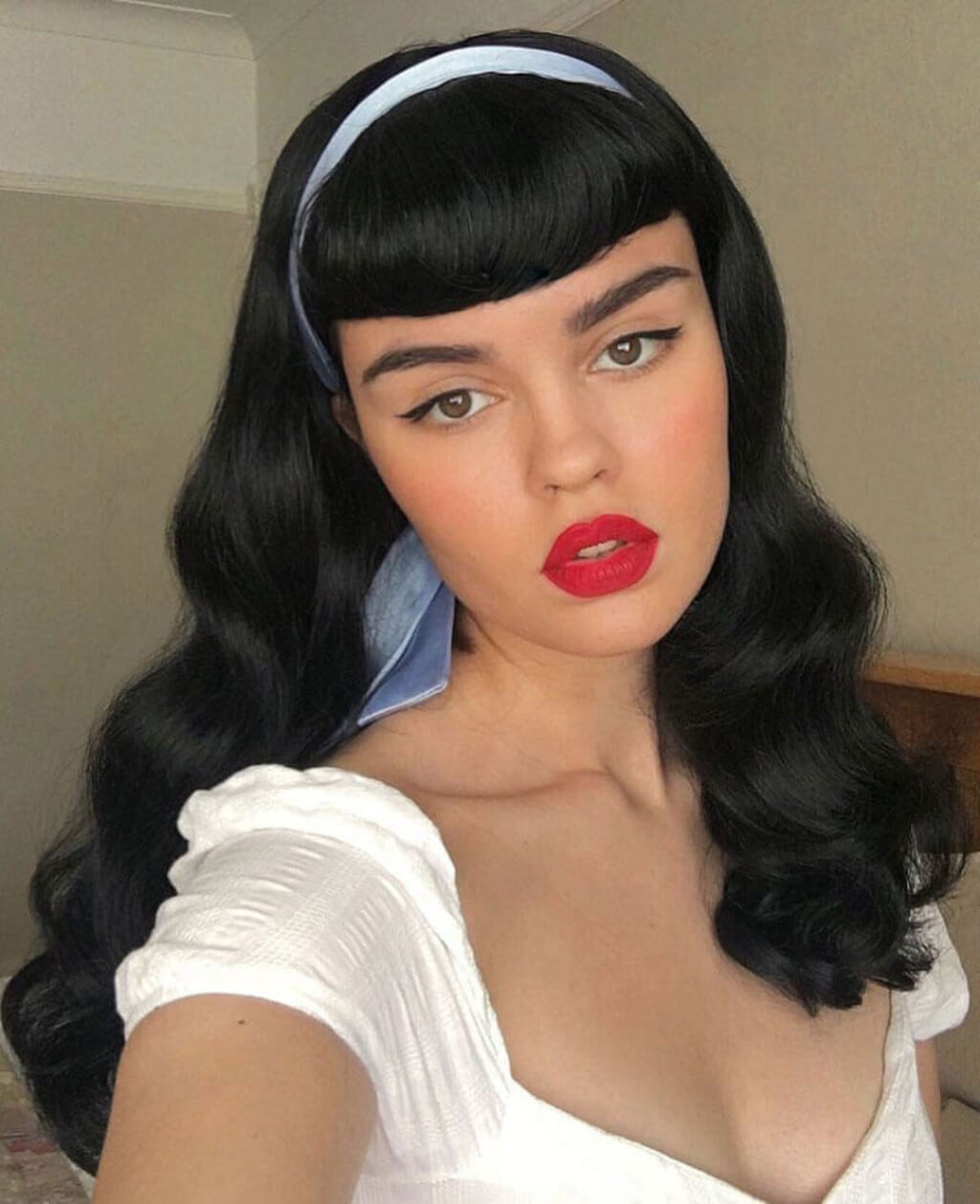 Woman with long black hair and red lipstick wearing a white top. Black 1950s pinup style wig, curled with short fringe: Milena