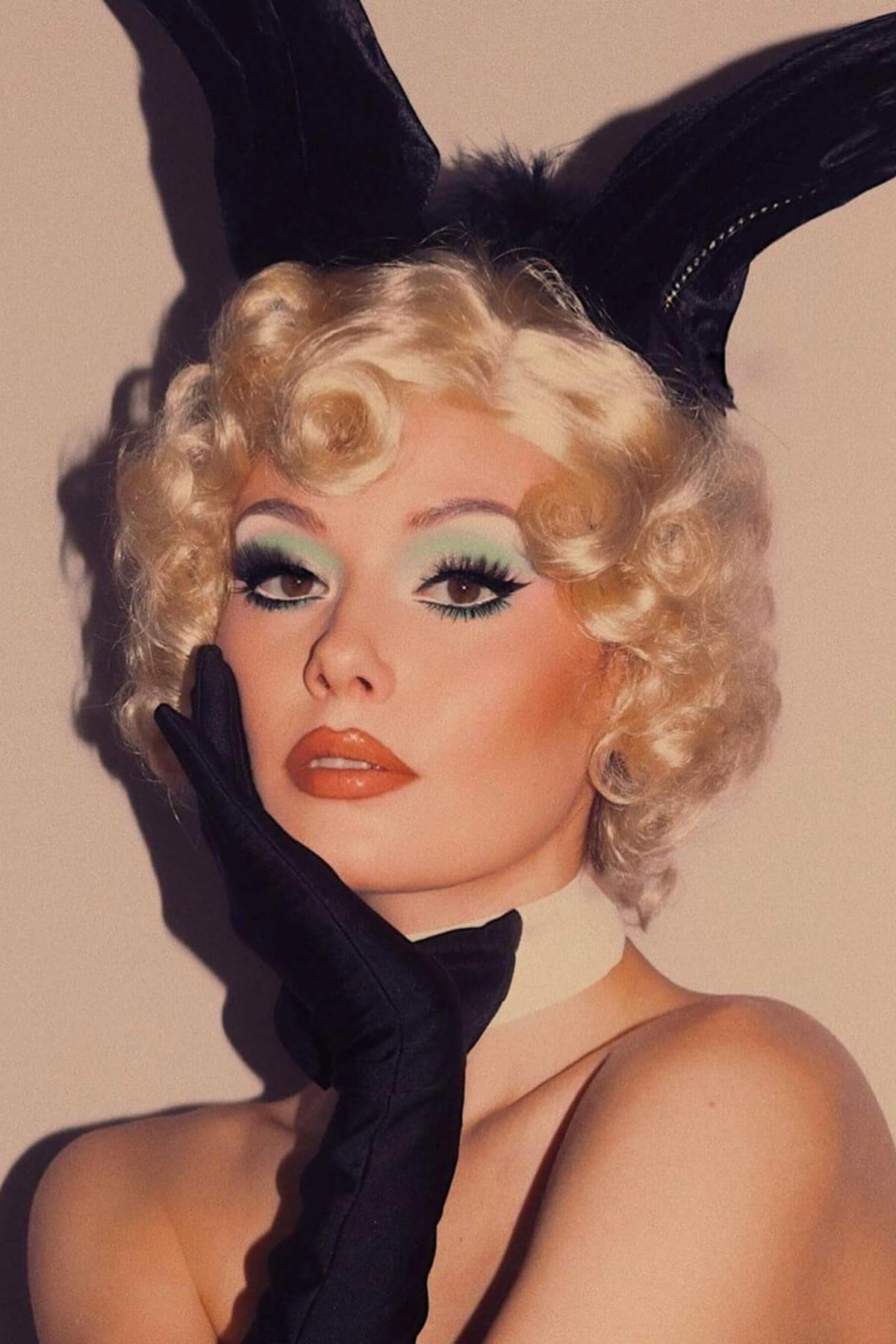Person with blonde wig, black gloves, and black headpiece against a beige background. Blonde 1920s 1930s style wig, short with finger waves: Diva champagne blonde 613