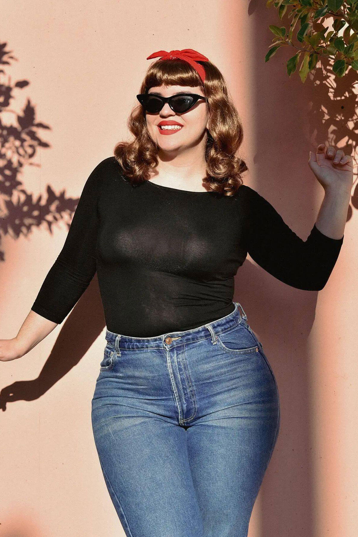 Woman wearing a black top and blue jeans with a red bandana against a pink wall. Brown pinup style wig, curled with short fringe: Carolina golden blonde 12-27